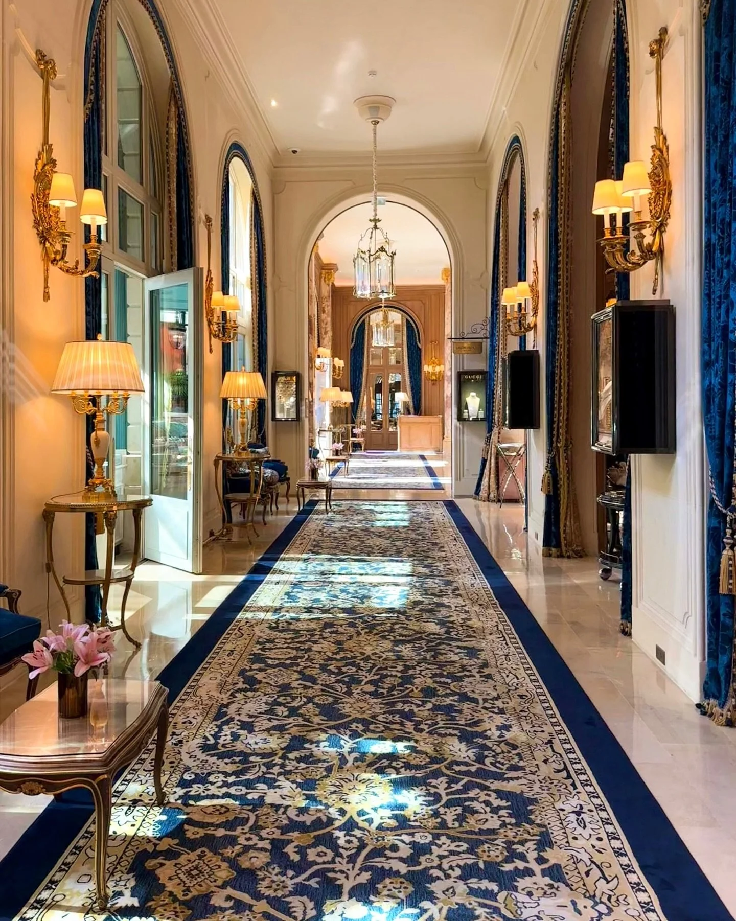 Un couloir élégant dans un hôtel ou un palais, avec un tapis bleu et doré, des lampes à appliques dorées, des rideaux bleus et des ornements dorés.