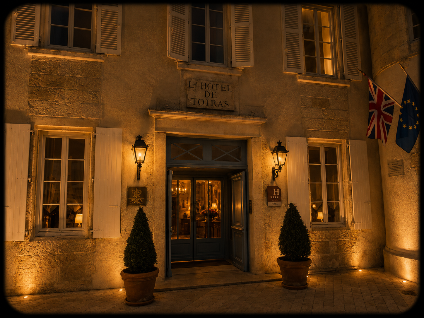 Facade du hôtel de Toiras éclairée la nuit avec deux drapeaux (Union Jack et Union Européenne) et deux lampes extérieures.