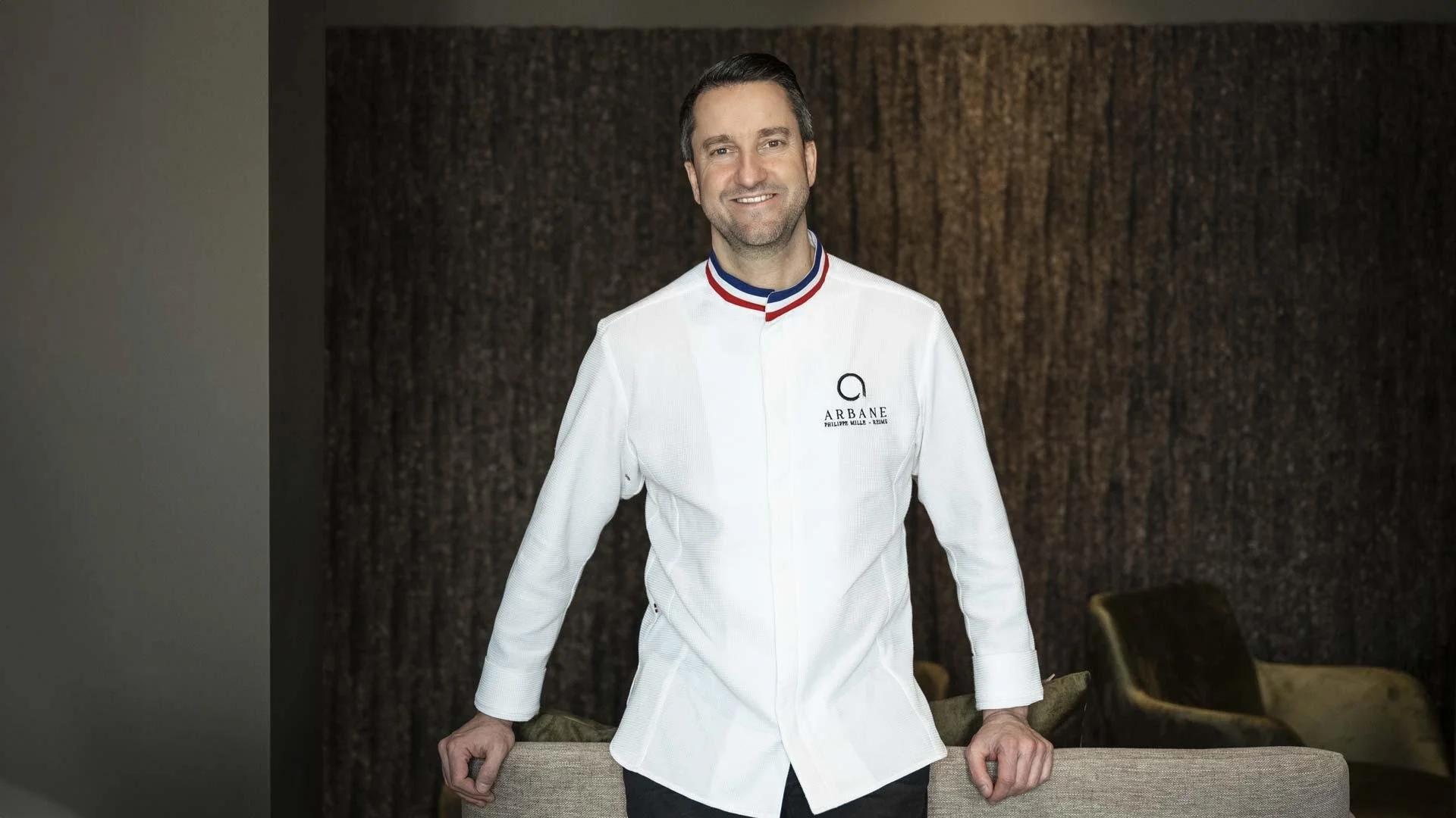 Homme souriant portant une veste de chef avec un logo ARBANE, debout derrière un canapé dans une pièce décorée.
