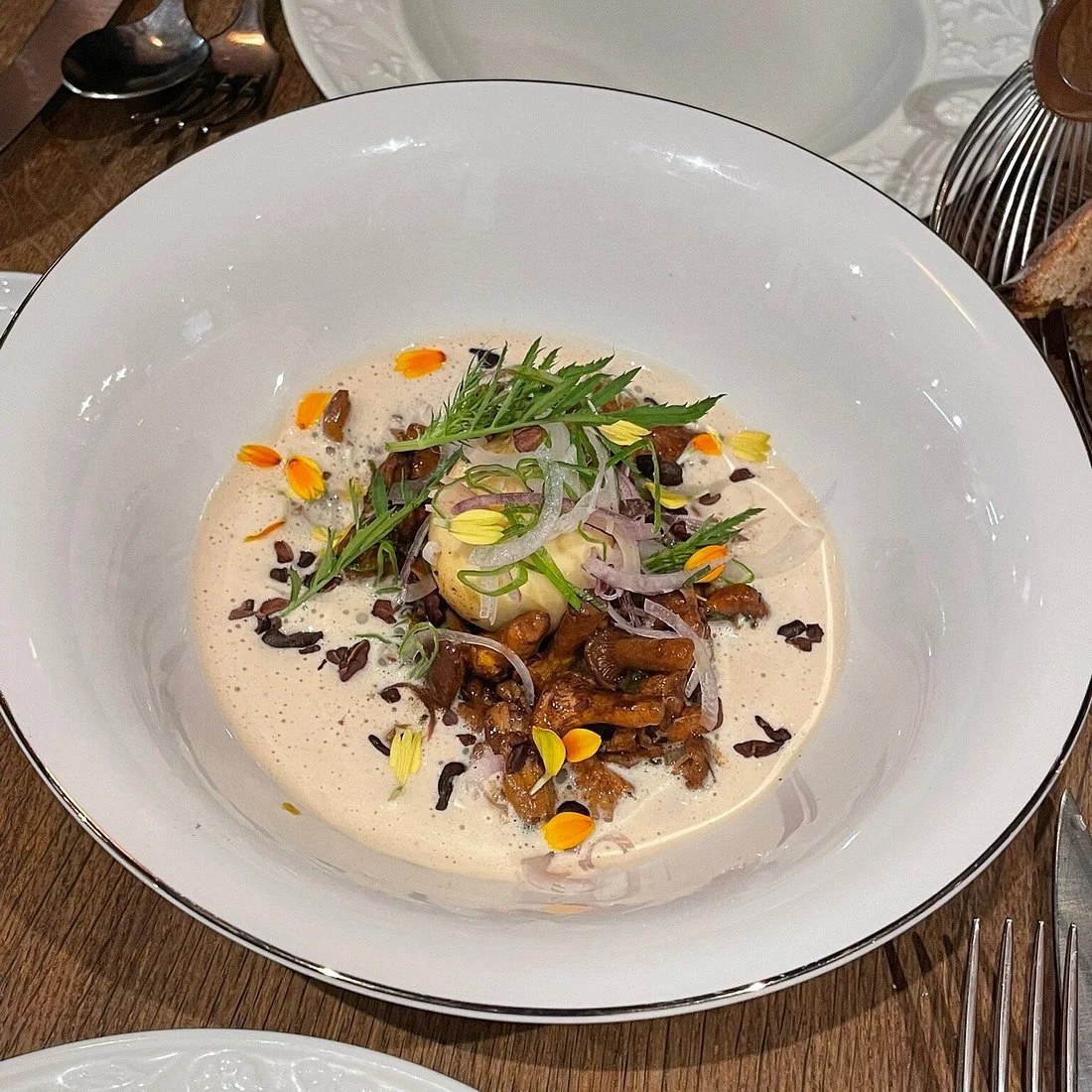 Un plat présenté dans un grand bol blanc contenant une crème ou une sauce claire, garnie de champignons mijotés, rondelles d'oignons, feuilles de verdure, petites fleurs jaunes et éclats de graines de tournesol.