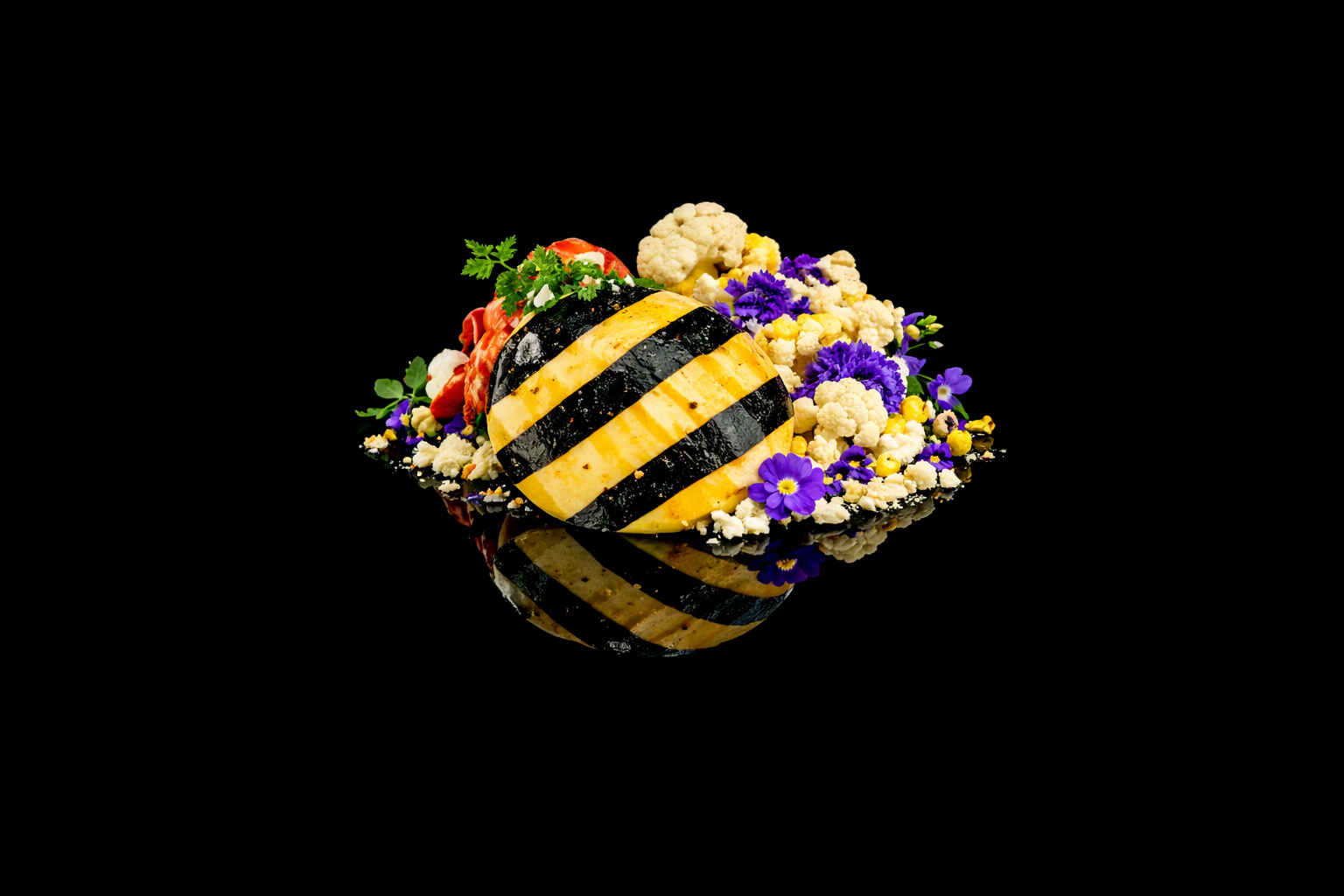Œuf de Pâques noir et jaune rayé entouré de fleurs violettes, blanches, et de légumes en miniature sur un fond noir reflété.