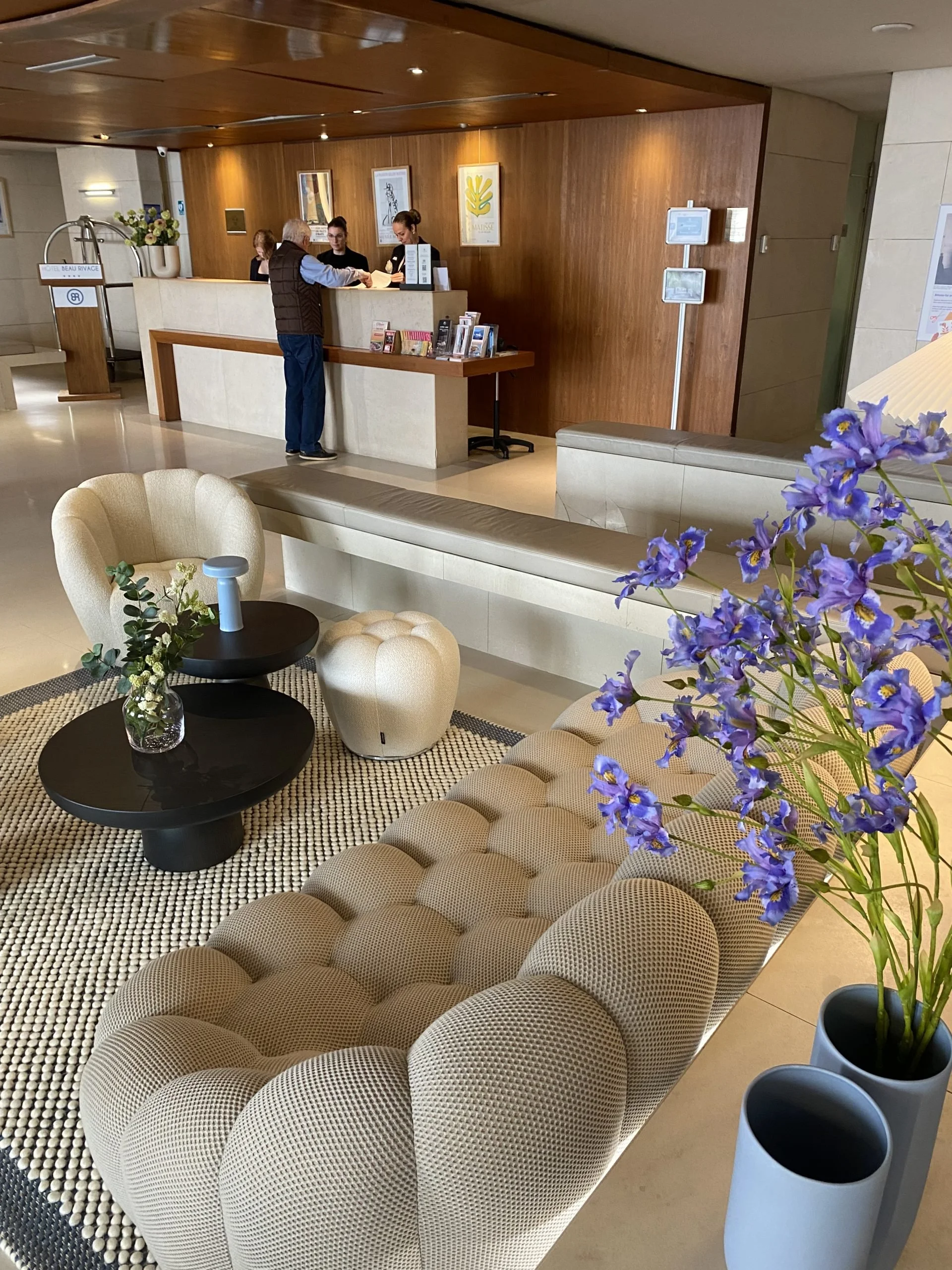 Un hall d'hôtel avec un coin lounge comprenant un canapé beige, une table basse noire avec une décoration florale, et des fauteuils en forme de pomme. À l'arrière, le comptoir de la réception avec du personnel accueillant un client.