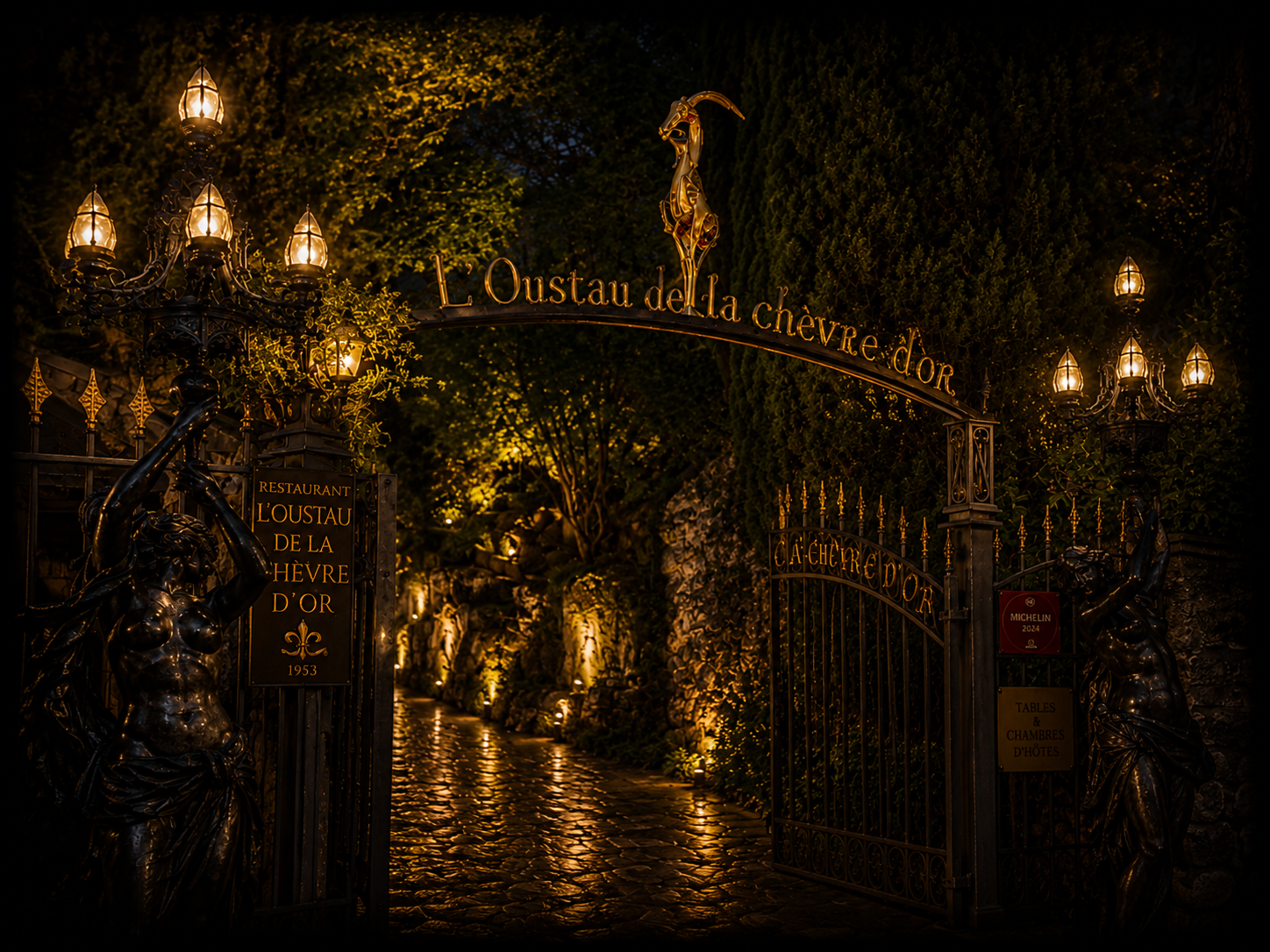 Une clôture ornementale en fer forgé orné de sculptures de femmes en bronze portant des lampes à gaz et une arche avec la signalisation 'L'Oustau de la chèvre d'or', entourée de végétation luxuriante illuminée la nuit.