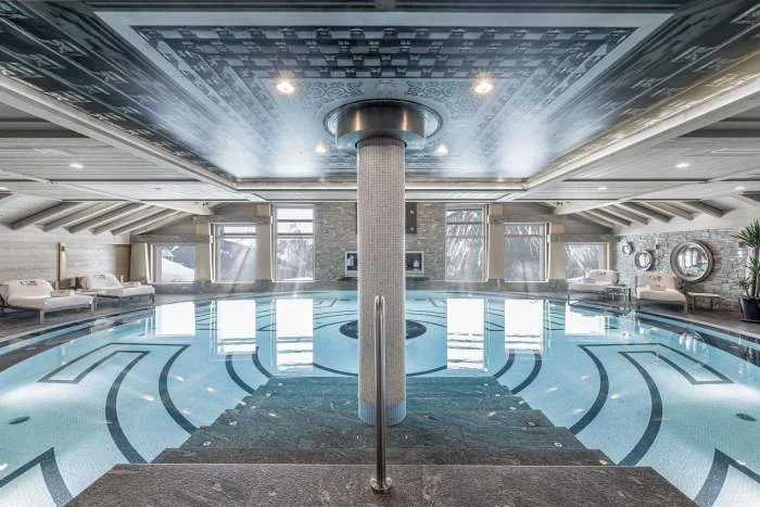 Piscine intérieure luxueuse avec colonnes, fenêtres, et mobilier élégant.