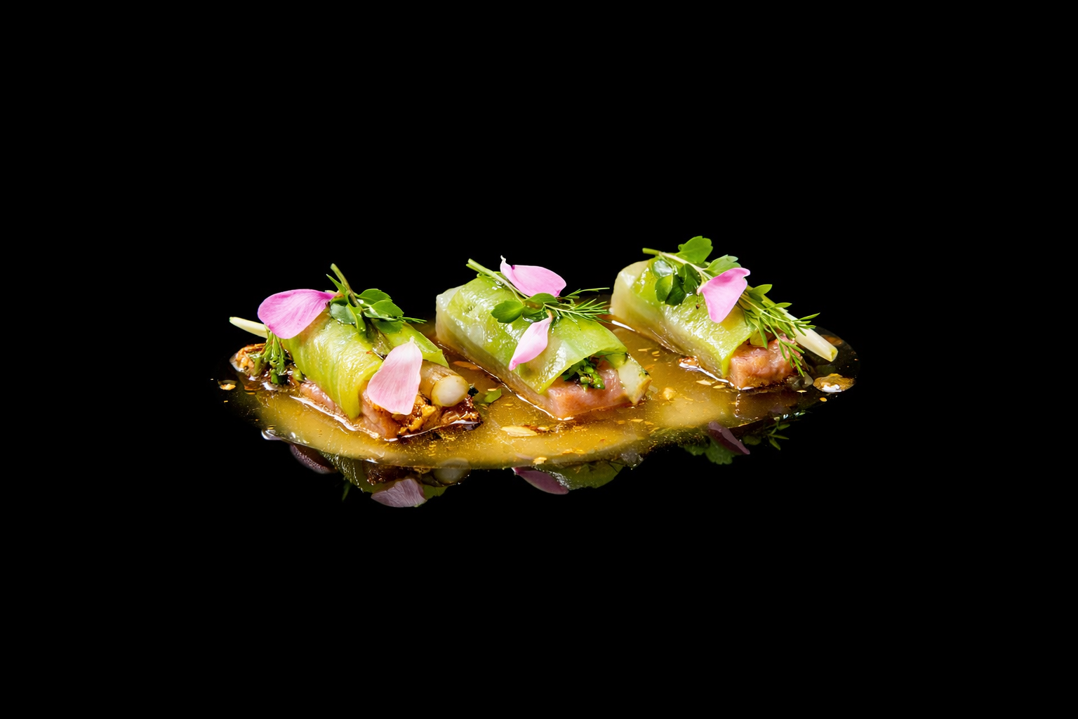 Trois amuse-bouches avec des rouleaux de concombre, garnis de plantes vertes et de pétales de fleurs, servis sur une sauce brillante, sur fond noir