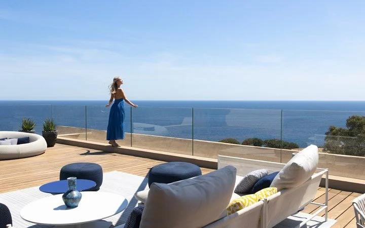 Femme en robe bleue regardant l'océan depuis une terrasse en bois équipée de mobilier de salon moderne.