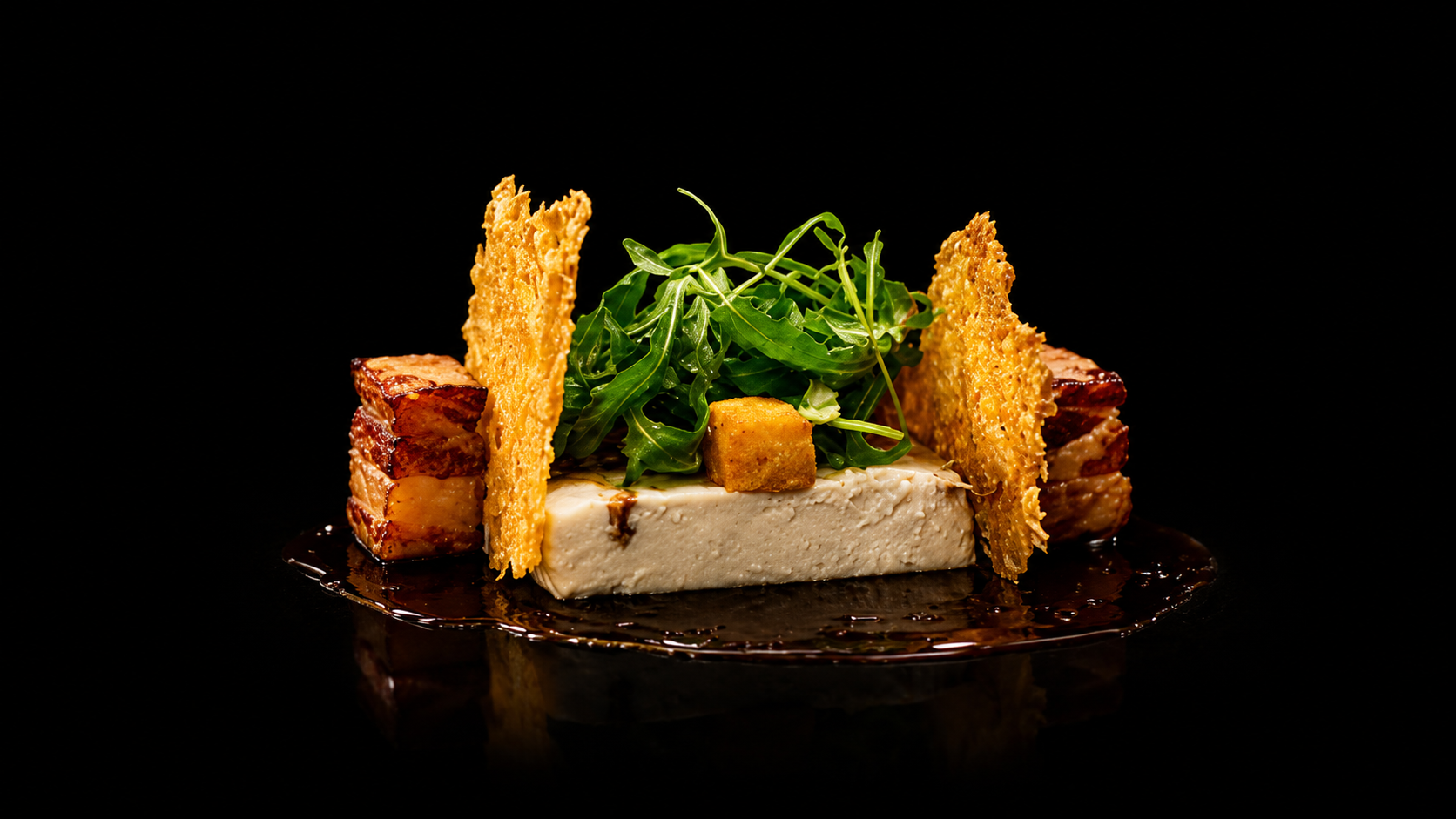 Assiette gastronomique avec du fromage, de la salade verte, des morceaux de bacon et des chips croustillantes sur un fond noir.