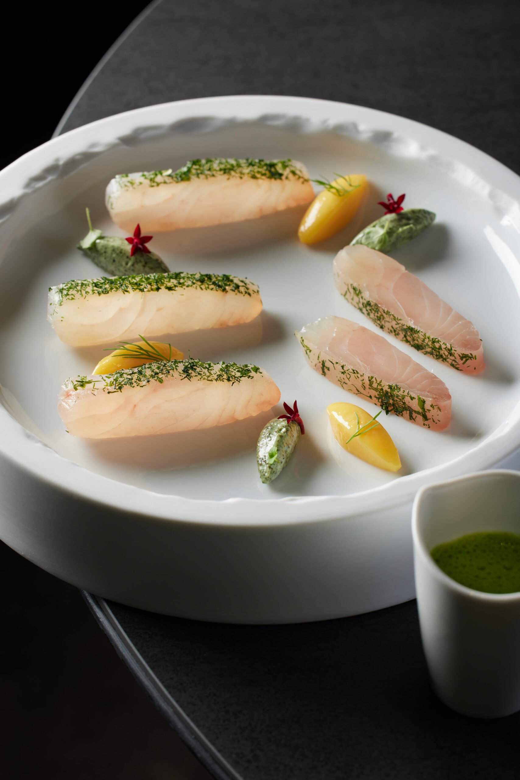 Assortiment de sashimis de poisson blanc, décoré de fines herbes, servi avec des tranches de citron, des sauces vertes, et des petites fleurs rouges, présenté sur une assiette blanche élégante.