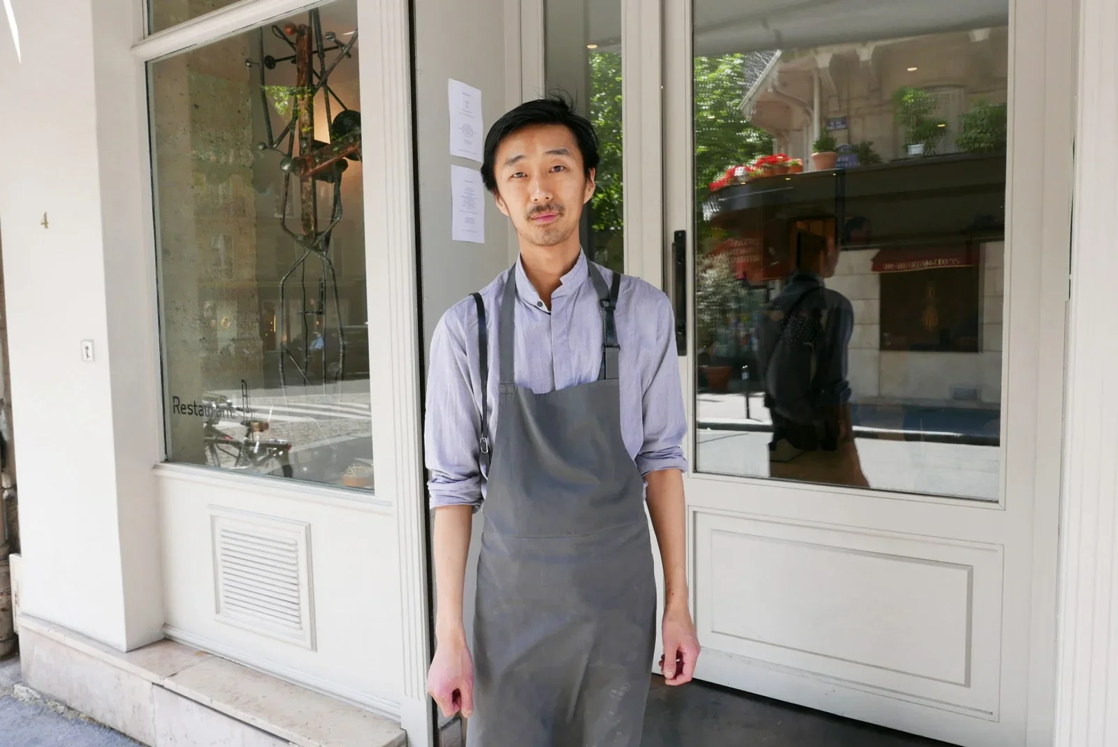 Un homme portant un tablier gris, debout devant une porte de restaurant, regarde l'appareil photo.