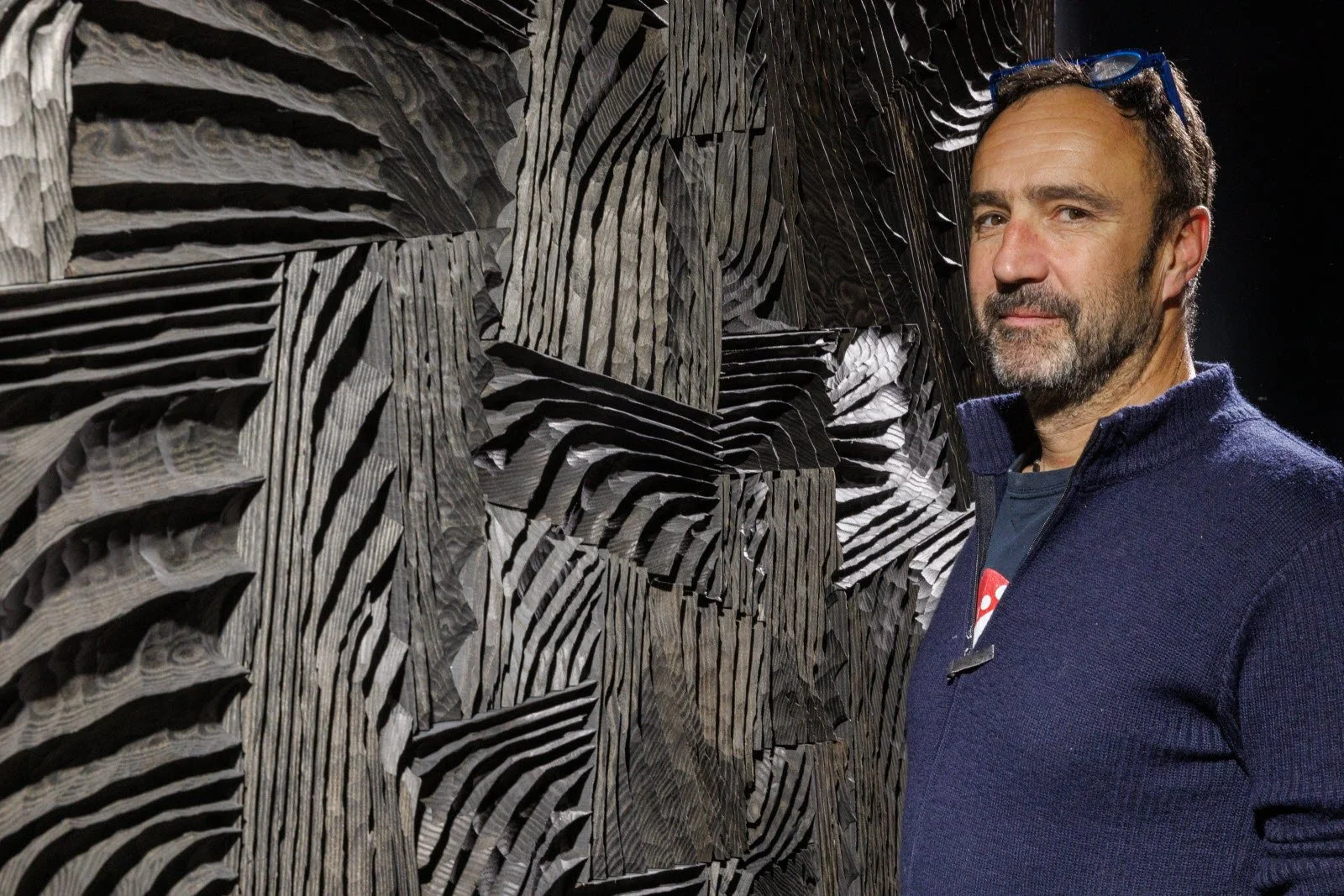 Un homme avec une barbe, portant des lunettes sur la tête, se tient devant une installation artistique en bois sculpté en motifs de lignes et de stries.