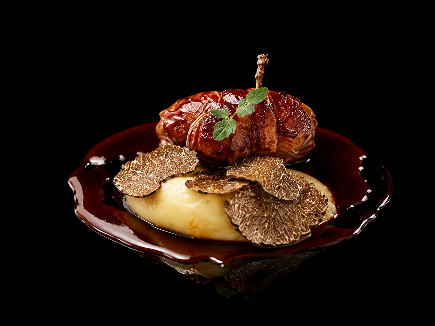 Plat gastronomique avec une portion de purée de pommes de terre garnie de truffes, une pièce de viande enroulée dans une sauce riche, décorée d'une herbe et d'une tranche de truffe séchée, présenté sur une assiette noire.