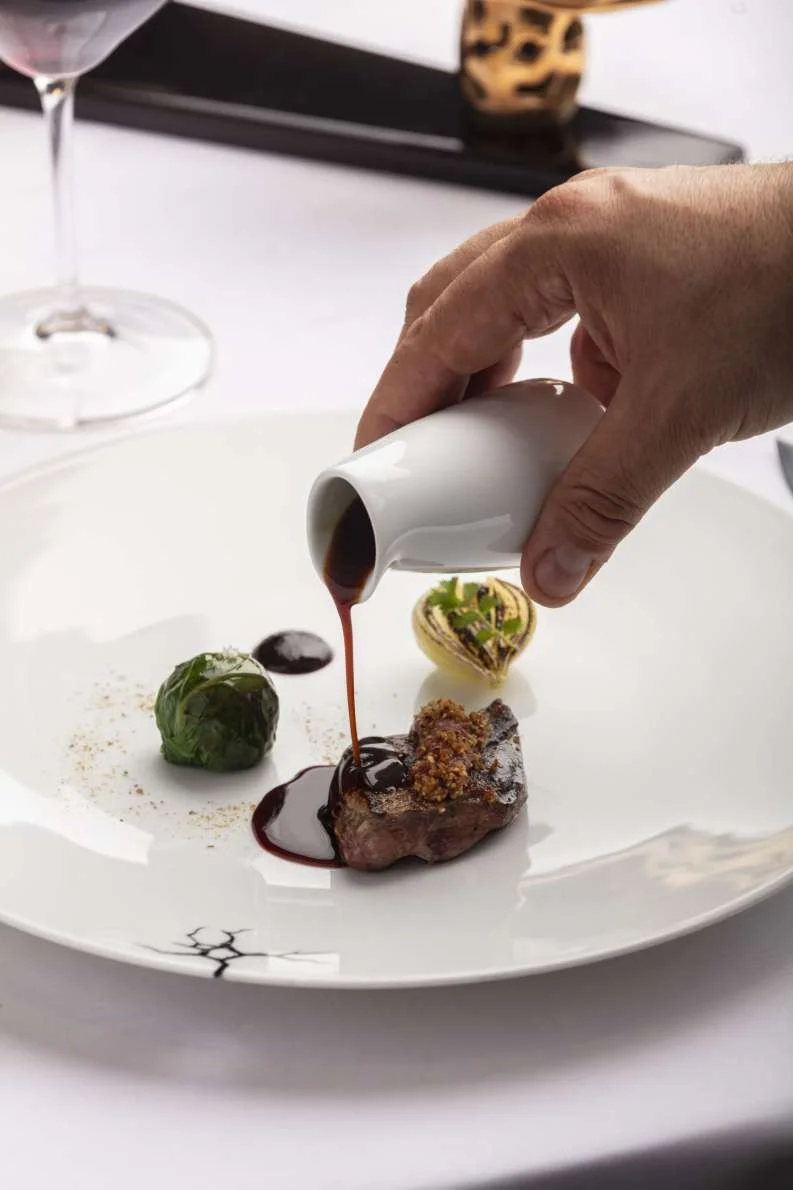 Une main verse une sauce foncée sur un plat gourmet avec une viande et des légumes.