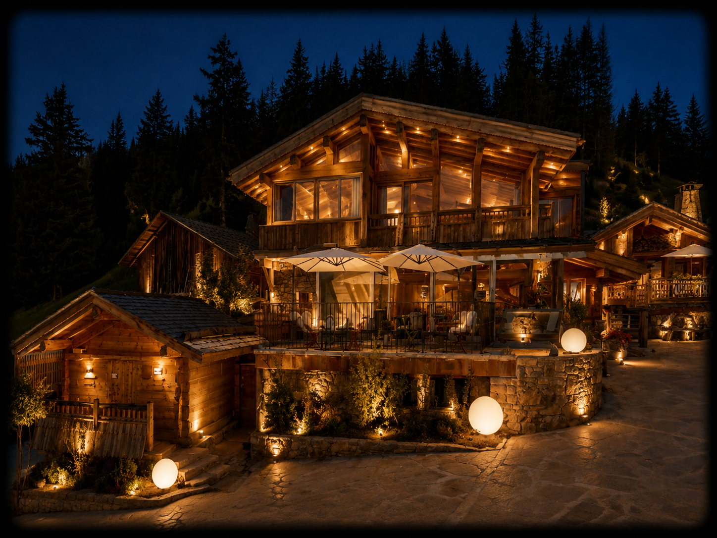 Une maison en bois éclairée la nuit, située en montagne avec un terrain pavé, avec un patio où se trouvent des parasols et des lampes rondes