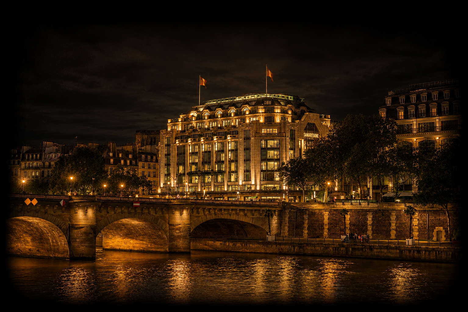 Bâtiment élégant illuminé la nuit, reflet dans la Seine, pont en pierre devant, arbres et lampadaires, ciel nuageux.