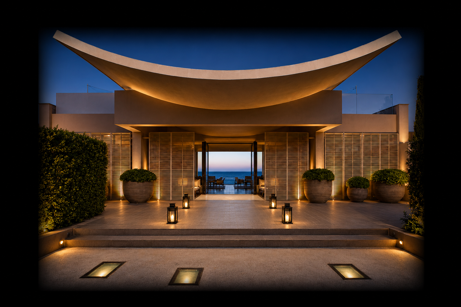 Bâtiment moderne avec un toit incurvé, éclairé par des lumières de jardin, donnant sur la mer au coucher du soleil, avec des plantes en pot et un espace d'entrée avec des lanternes.
