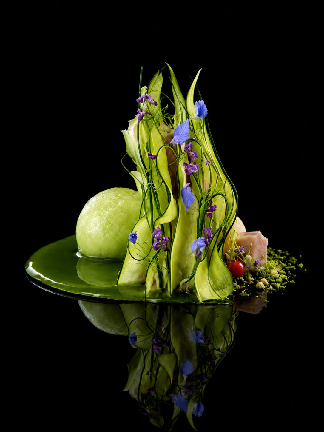Présentation gastronomique avec des légumes étagés, décoré de fleurs violettes et de graines, sur un fond noir reflété sur une surface brillante.