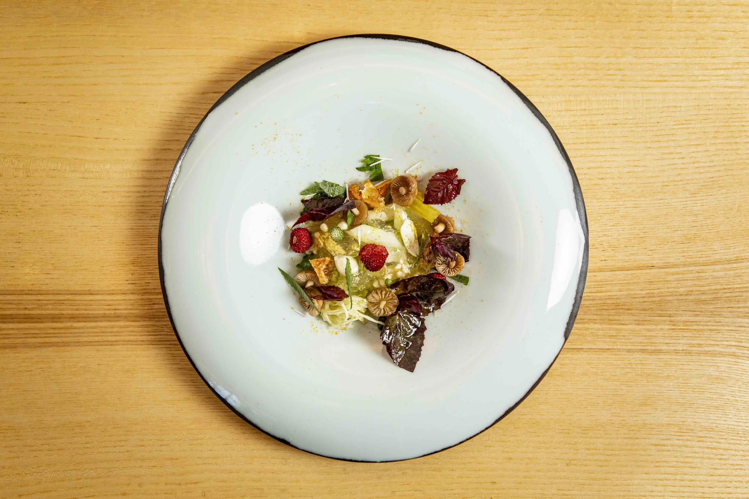 Assiette présentée avec un dessert ou une salade composée de fruits, de feuilles de salade, de champignons et de divers éléments colorés. Plateau en bois clair.