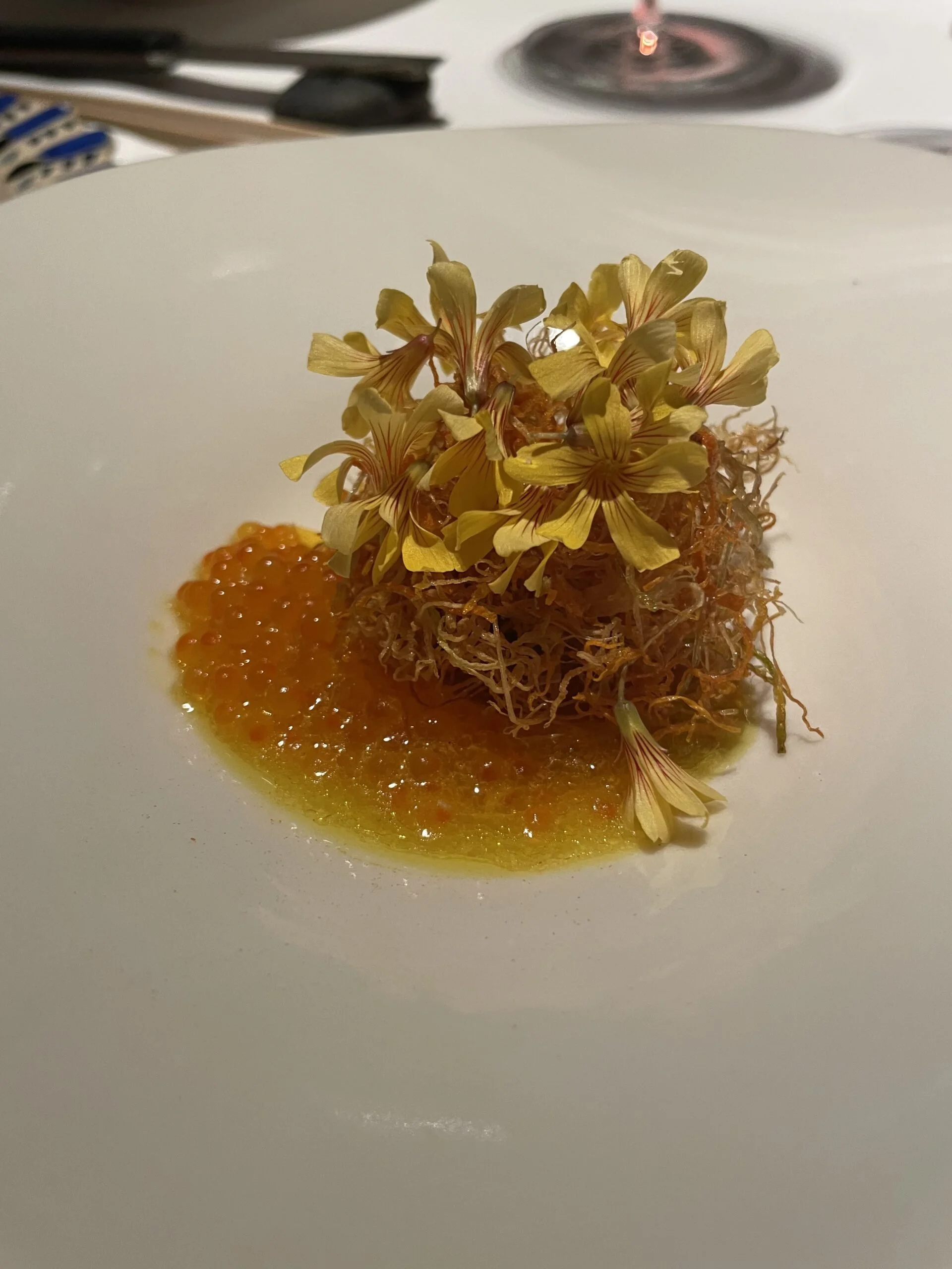 Plat de cuisine sophistiquée avec un monticule de garniture délicate, fleurs comestibles jaunes sur le dessus, accompagnée d'une sauce orange translucide sur une assiette blanche.