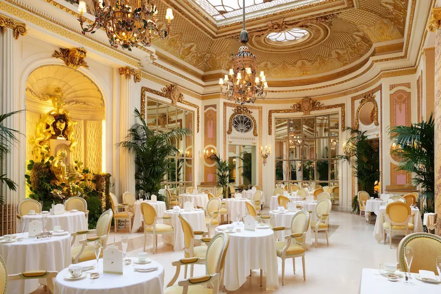 Salle de réception ornée de dorures, chandeliers en cristal, grandes plantes vertes, tables rondes avec nappes blanches et chaises élégantes, décoration baroque et lumière chaleureuse.