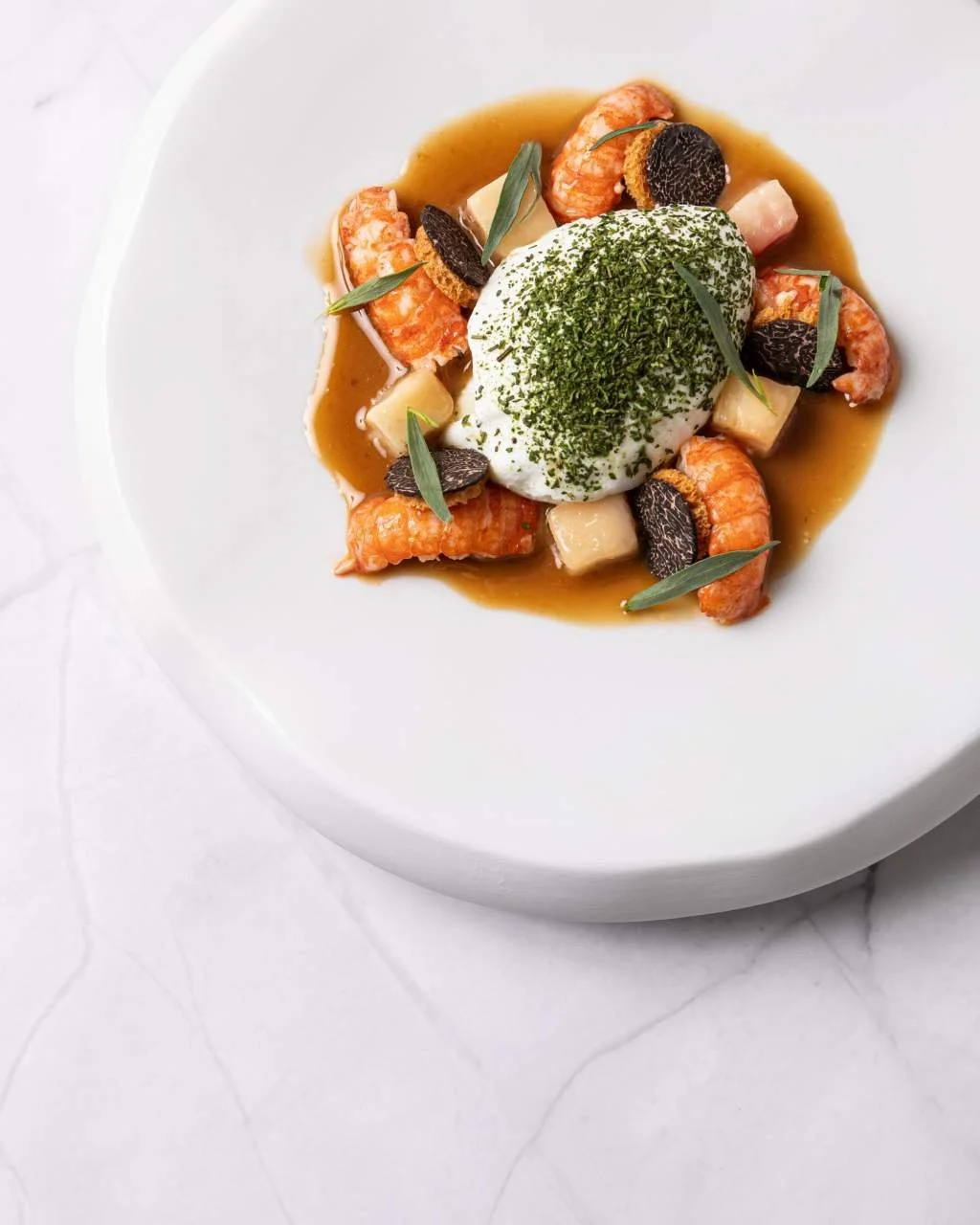 Plat gastronomique avec crevettes, œuf mollet, truffes, herbes aromatiques, sauce, et cubes de légumes, présenté dans une assiette blanche.