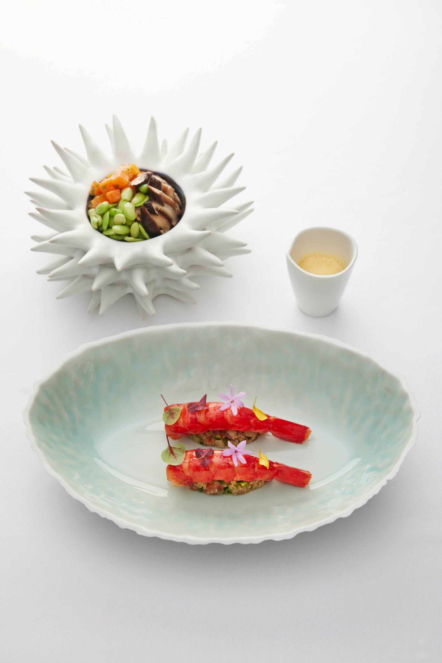 Deux plats de cuisine fine, un avec deux morceaux de homard garnis de fleurs comestibles, l'autre avec un bol sculpté en forme de fleur contenant un mélange de légumes et de viande, accompagnés d'un petit récipient contenant une poudre ou une épice.