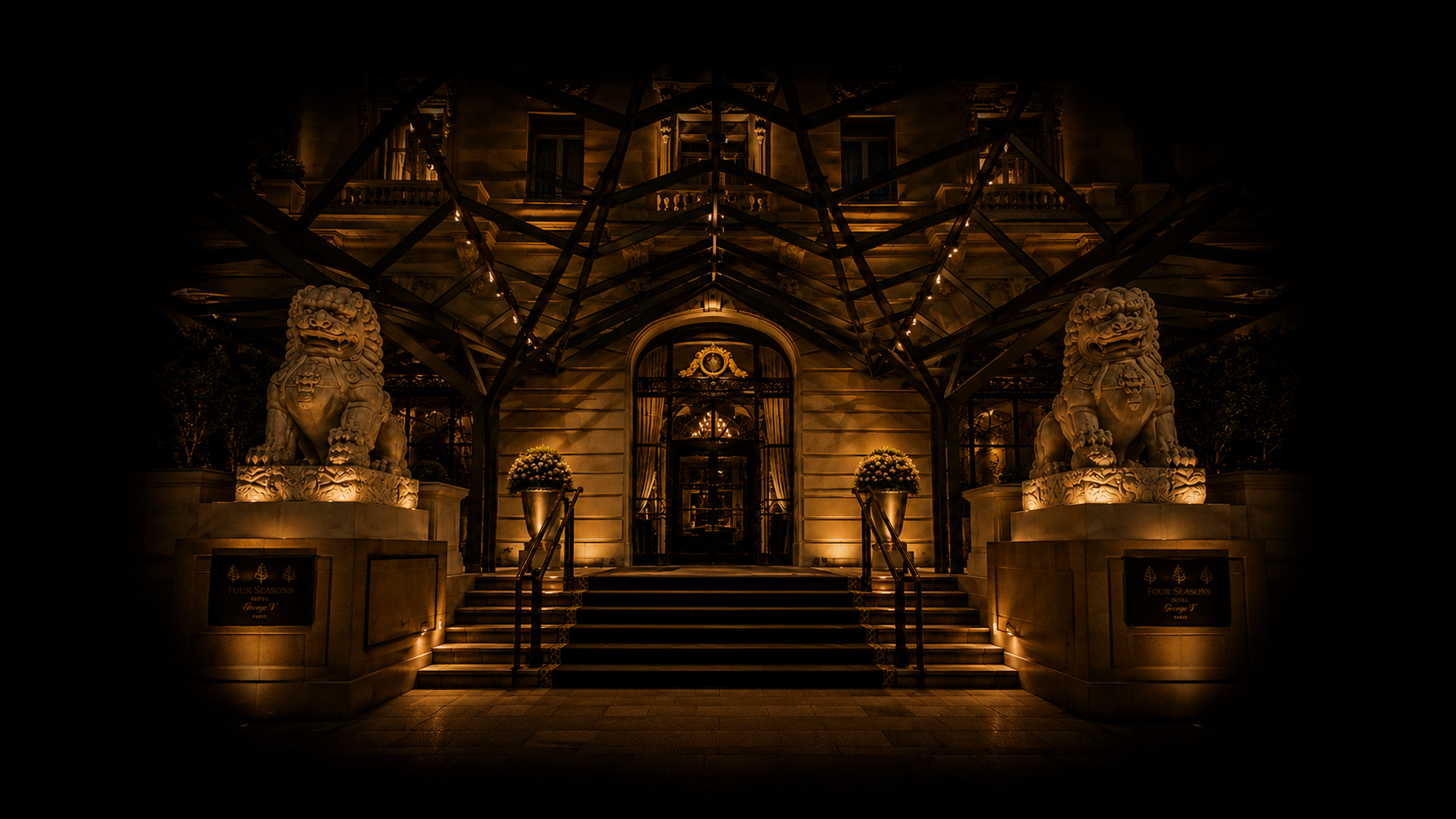 Entrée d'un hôtel de luxe avec deux lions en pierre illuminés la nuit, des escaliers, des plants de fleurs en pots, un portail en fer forgé et une structure métallique au-dessus, éclairés par un éclairage chaud.