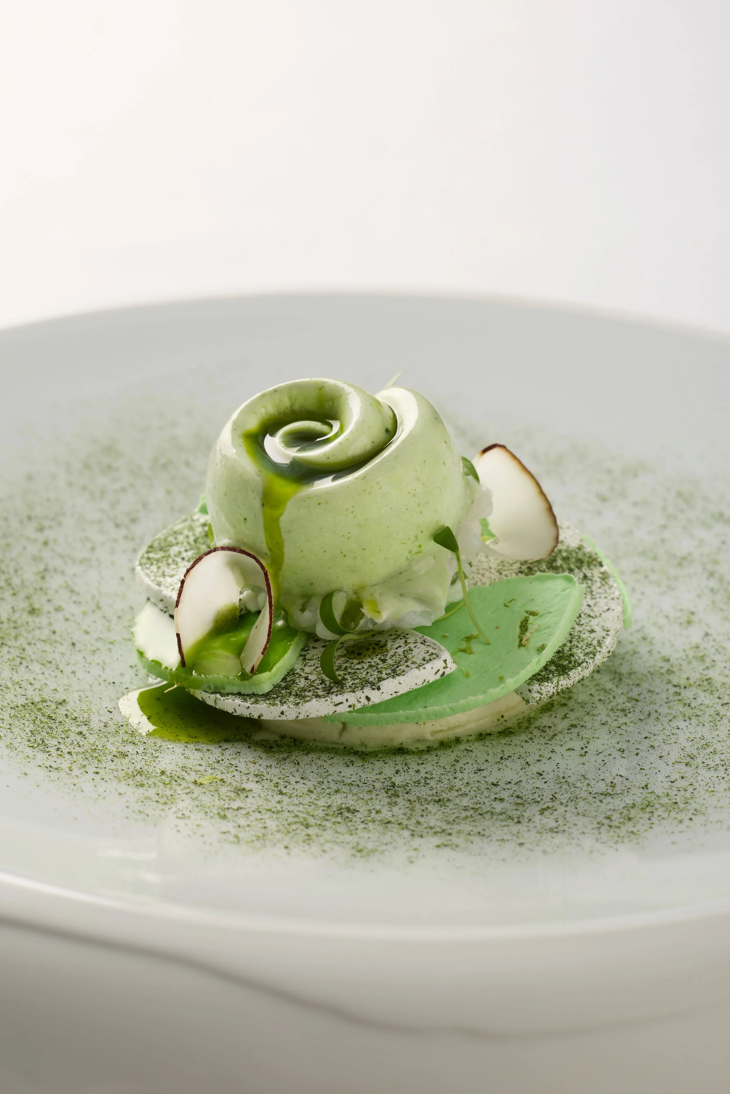 Dessert en forme de rose verte avec des tranches de noix de coco, des feuilles de menthe et une poudre verte sur une assiette blanche.
