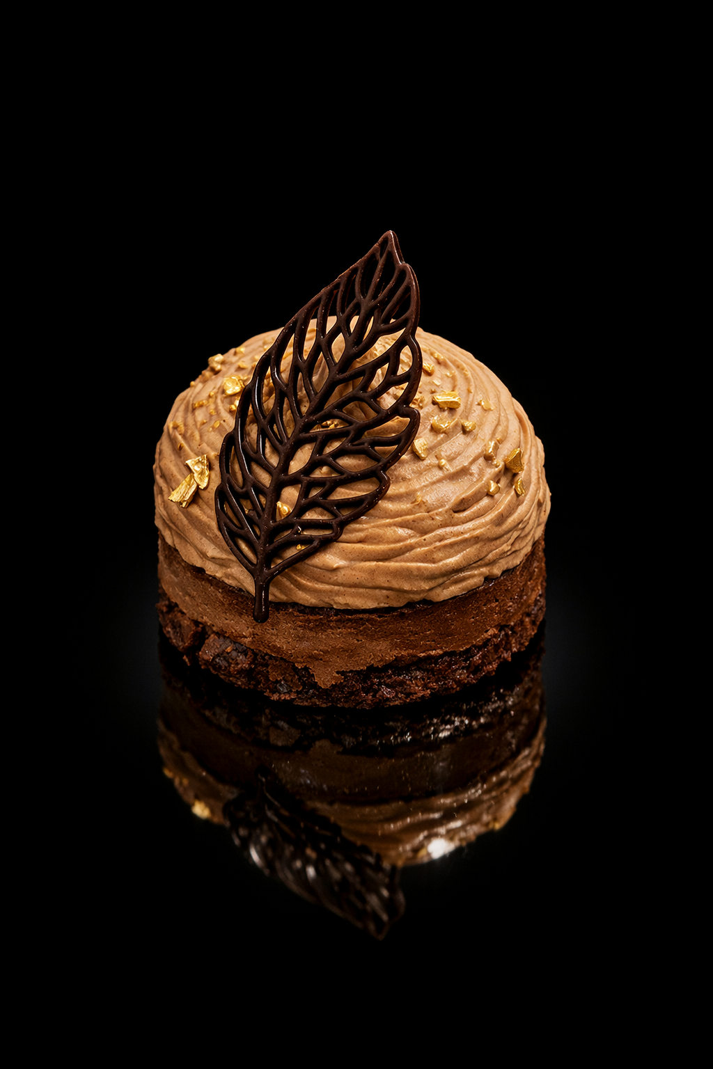 Petite pâtisserie au chocolat décorée d'une fine feuille en chocolat et de petits croquants dorés, avec un glaçage crémeux et une base croustillante, sur fond noir.
