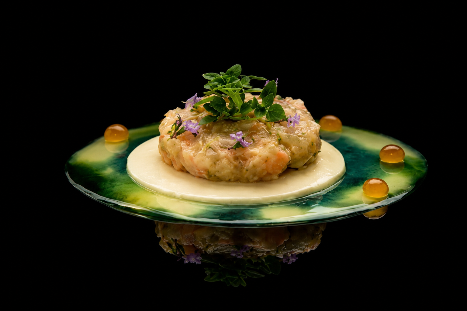 Tartare de poisson garni de microgreens et de petites fleurs violettes, présenté sur une assiette artistique avec des perles d'un liquide orange et une sauce verte sur fond noir.