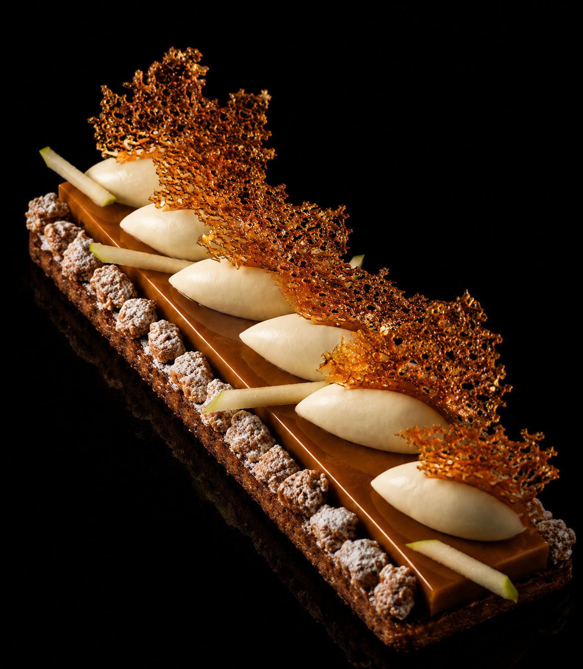 Un dessert sophistiqué avec une base en chocolat, décoré de crumbles, de tranches de pomme verte, de poires en pâte, et une fine plaque de caramel avec une feuille de caramel croquante en forme de dentelle.
