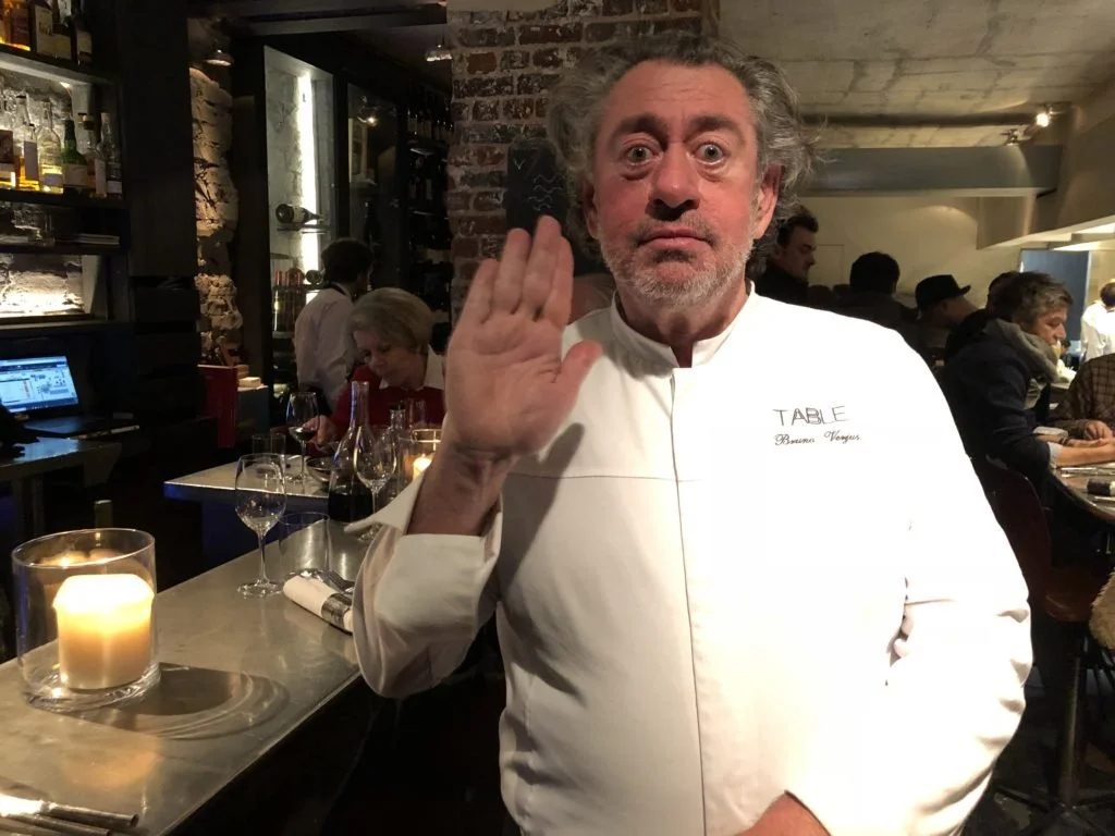 Chef en uniforme blanc saluant dans un restaurant chic avec de la nourriture et des boissons sur la table, et des clients assis en arrière-plan.