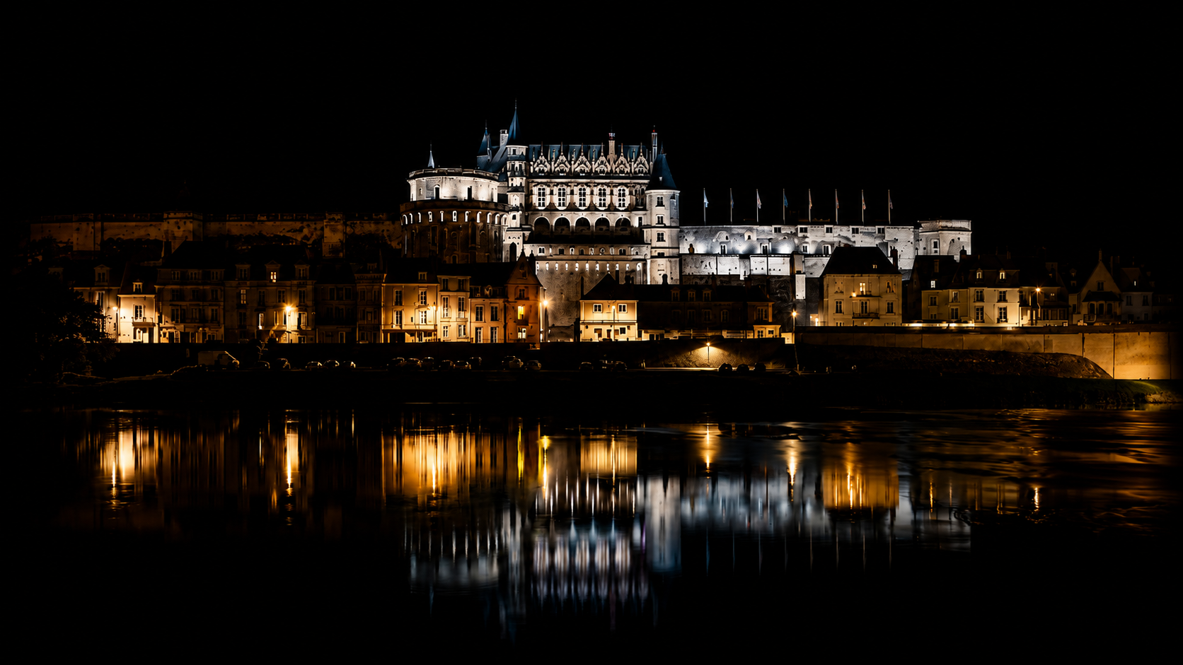 Château illuminé la nuit, reflet dans l'eau, bâtiments résidentiels en dessous.