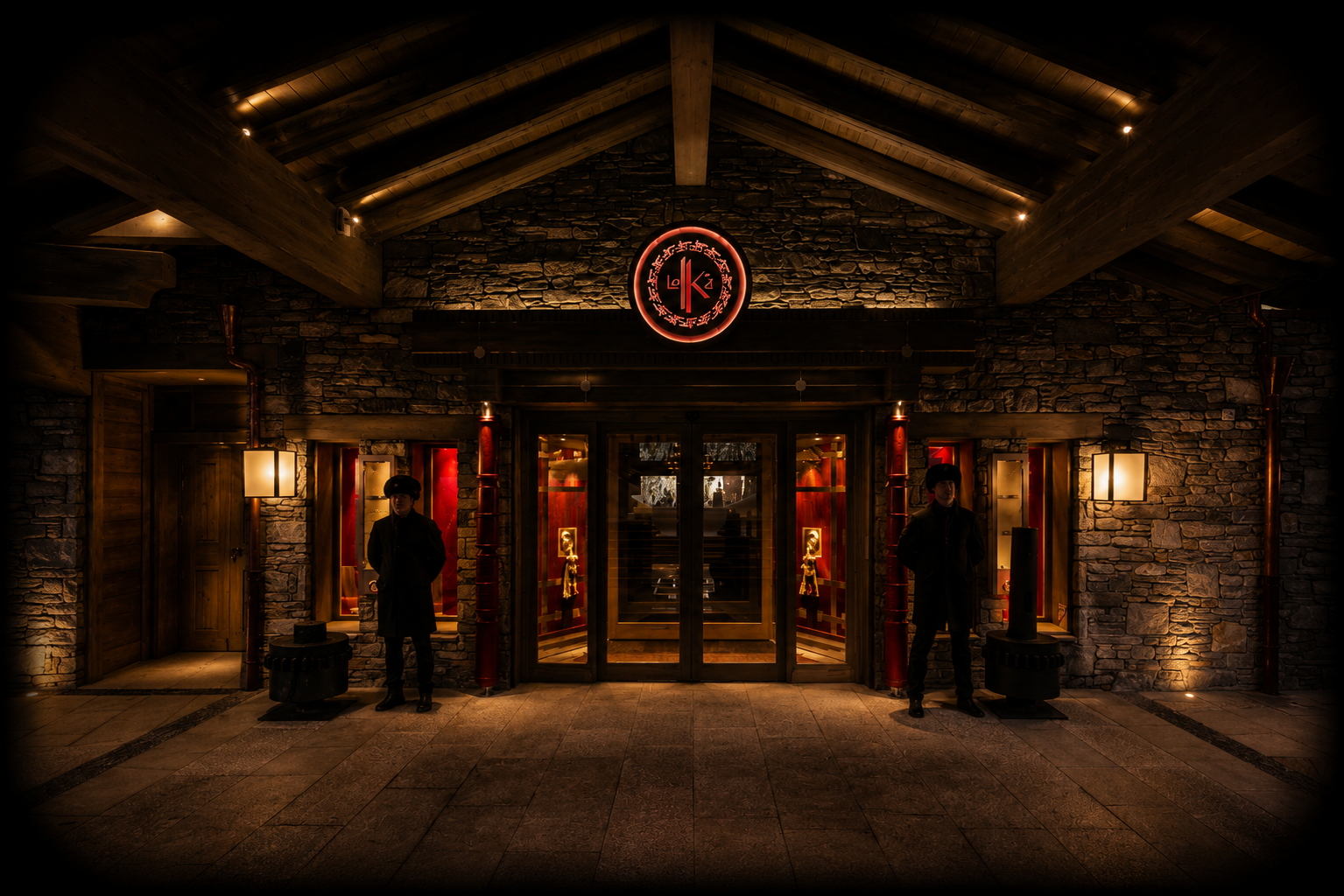 Entrée d'un restaurant avec une façade en pierre et un toit en bois, éclairée par des lampes, deux personnes vêtues de noir en garde à l'entrée, et un logo lumineux avec des symboles au-dessus de la porte.