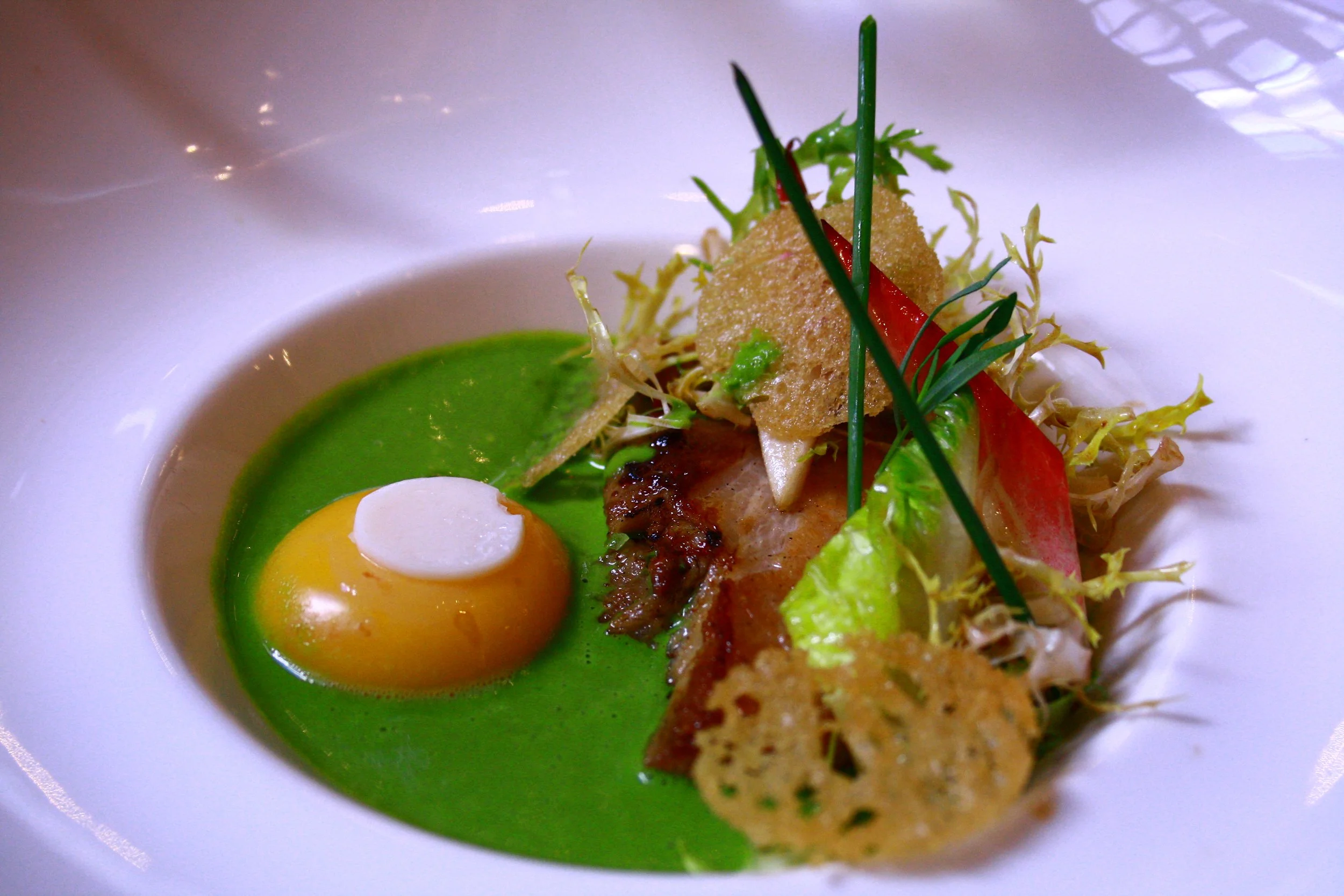 Assiette de nourriture gastronomique avec un œuf coulant, purée verte, morceaux de viande, chips, légumes, et herbes fraîches.
