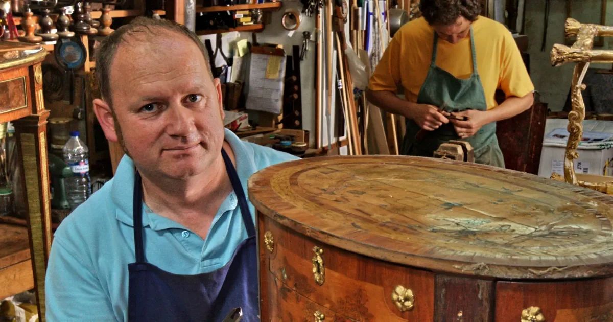 Un homme dans un atelier de restauration de meubles, manipulant une table ancienne en bois ornée de dorures. En arrière-plan, une femme travaille avec des outils.