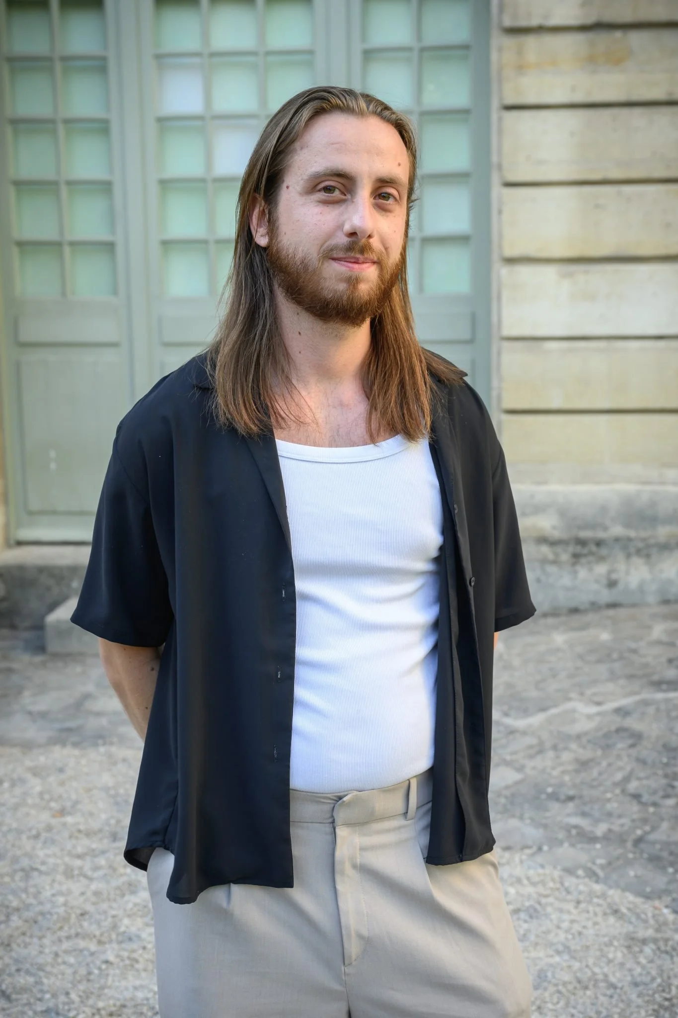 Jeune homme avec long cheveux, barbe, porte un t-shirt blanc, chemise noire ouverte et pantalon beige, debout devant un bâtiment en pierre avec fenêtres à carreaux.