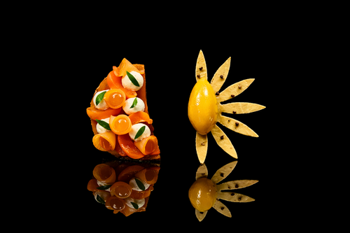 Deux plats artistiques de nourriture présentés sur un fond noir. La première est une composition colorée avec du saumon, des œufs de poissons, et des feuilles vertes. La seconde ressemble à un fruit jaune décoré avec des tranches de pomme disposées autour comme des pétales.