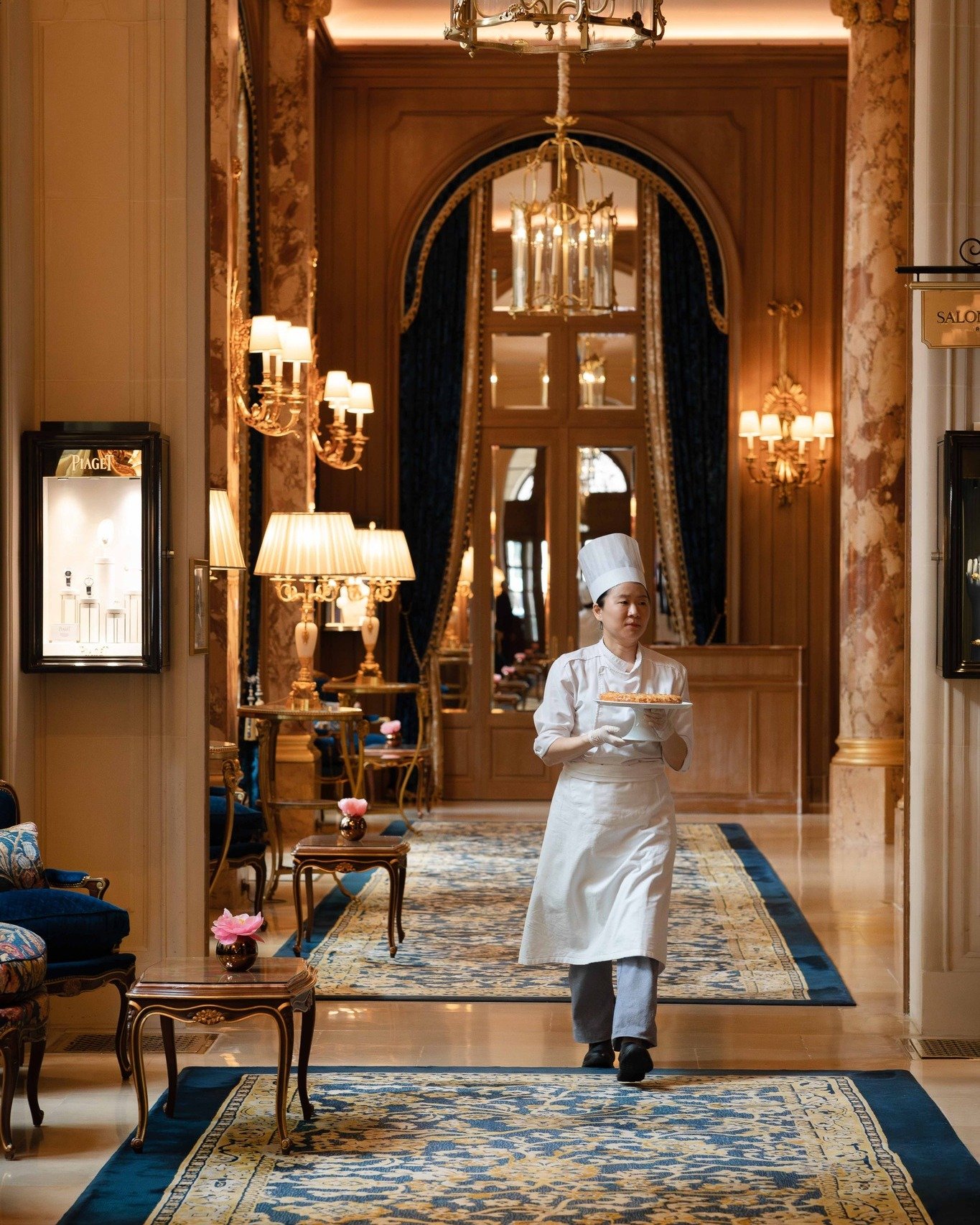 Une serveuse en uniforme blanc portant un plat de nourriture dans un élégant hôtel ou restaurant, avec un décor sophistiqué comprenant des tapisseries, des lampes et des meubles en bois.