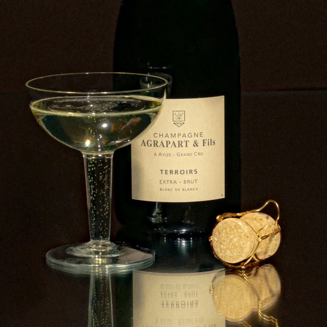 Une bouteille de champagne Agrapart & Fils, un verre de champagne remplie, et un morceau de champagne en bouchon à champagne avec un anneau en or posé sur une surface reflective.