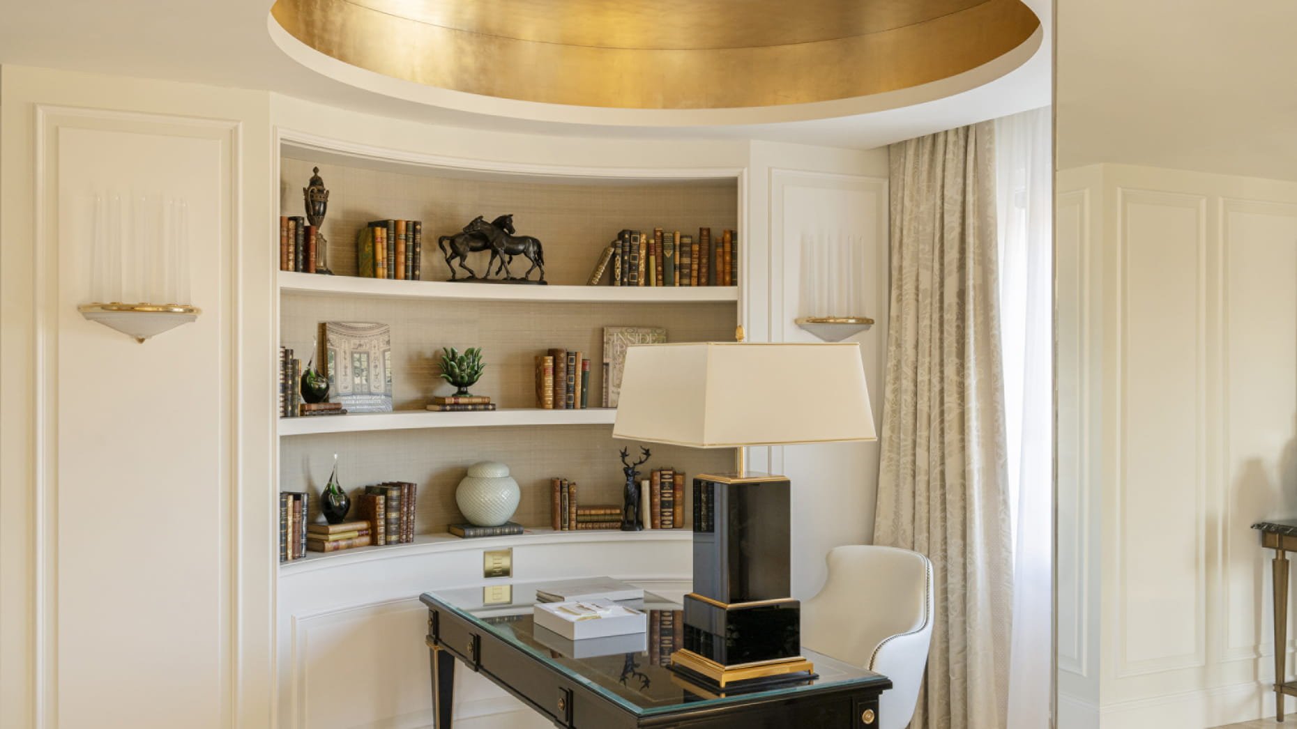 Espace de lecture élégant avec une cheminée, étagères remplies de livres, décorations en céramique et figurines, grande lampe de table et fauteuil blanc avec bras selon une ambiance sophistiquée.