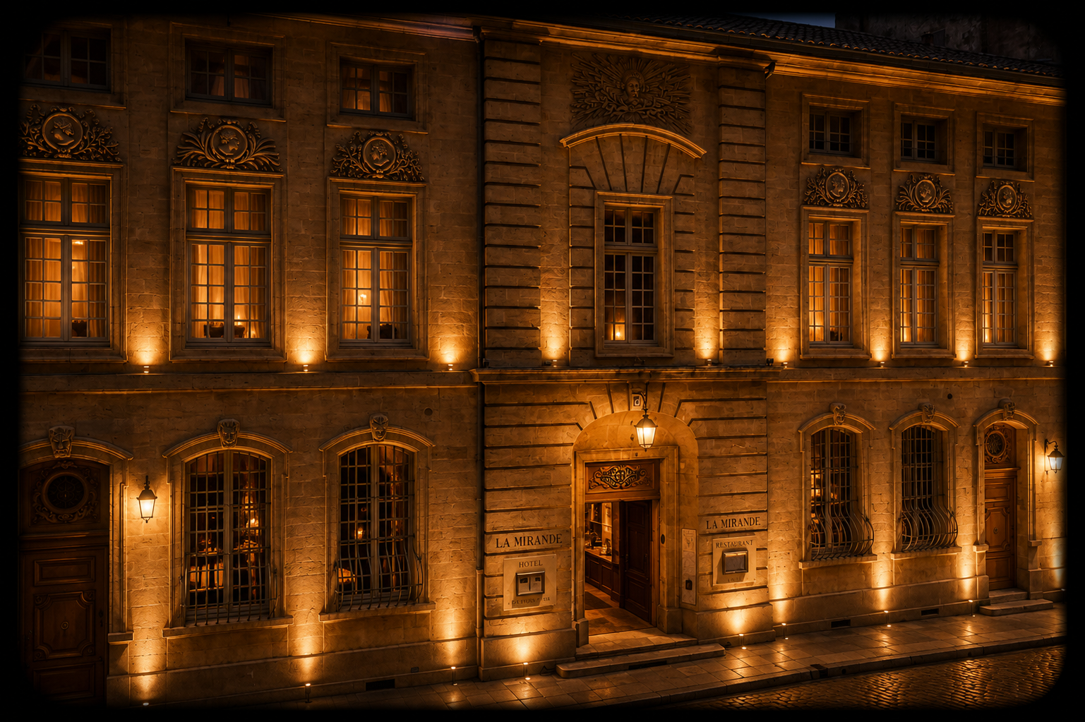 Un bâtiment historique illuminé la nuit, connu sous le nom de La Mirande, avec des fenêtres éclairées, des lampes extérieures et une façade en pierre ornée.