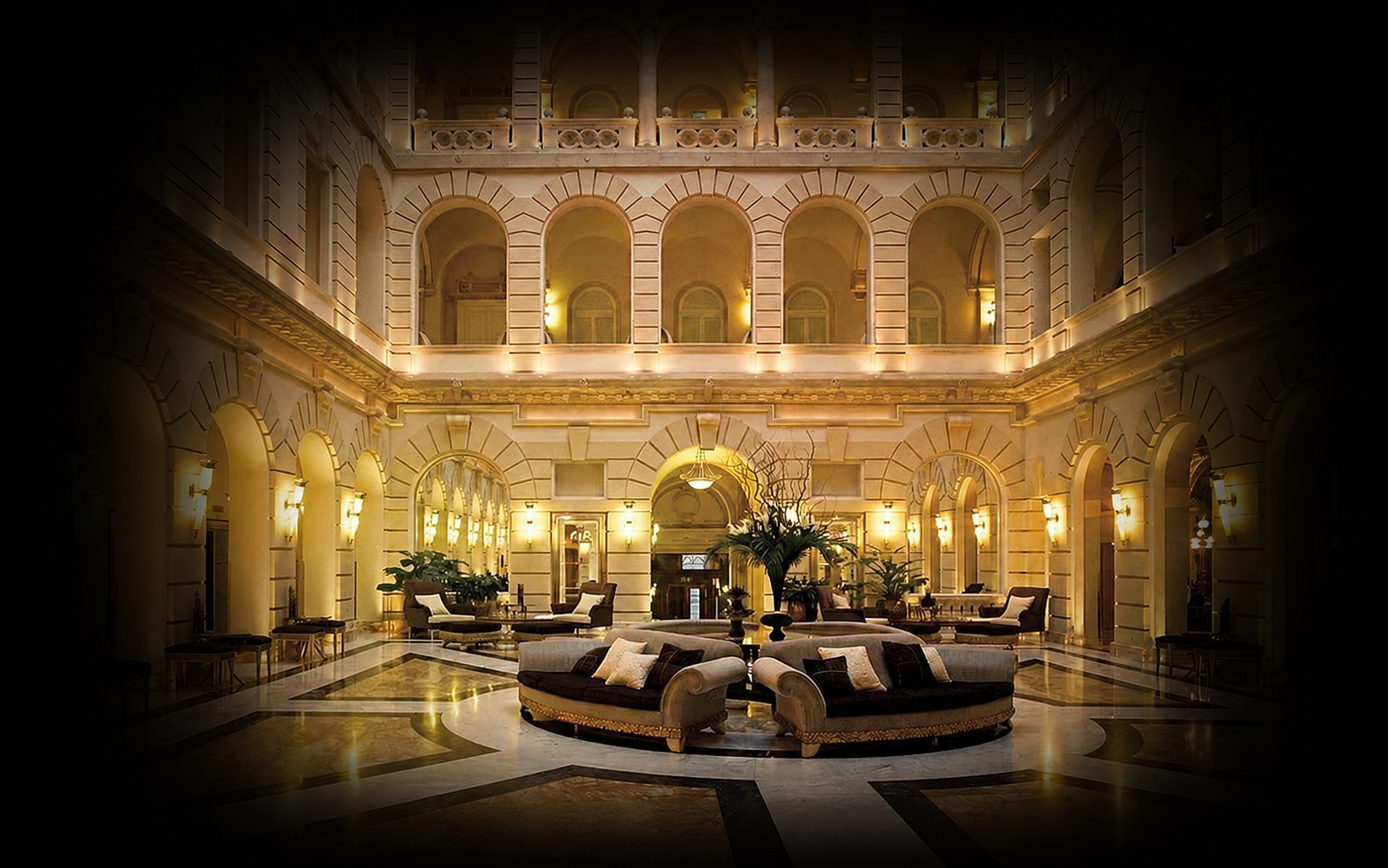 Vue intérieure d'un hall de luxe avec des canapés, des tables basses, des plantes et un décor élégant en avant-plan, avec un grand escalier et des fenêtres à arches à l'étage, éclairés par des lampes dans un bâtiment sophistiqué.
