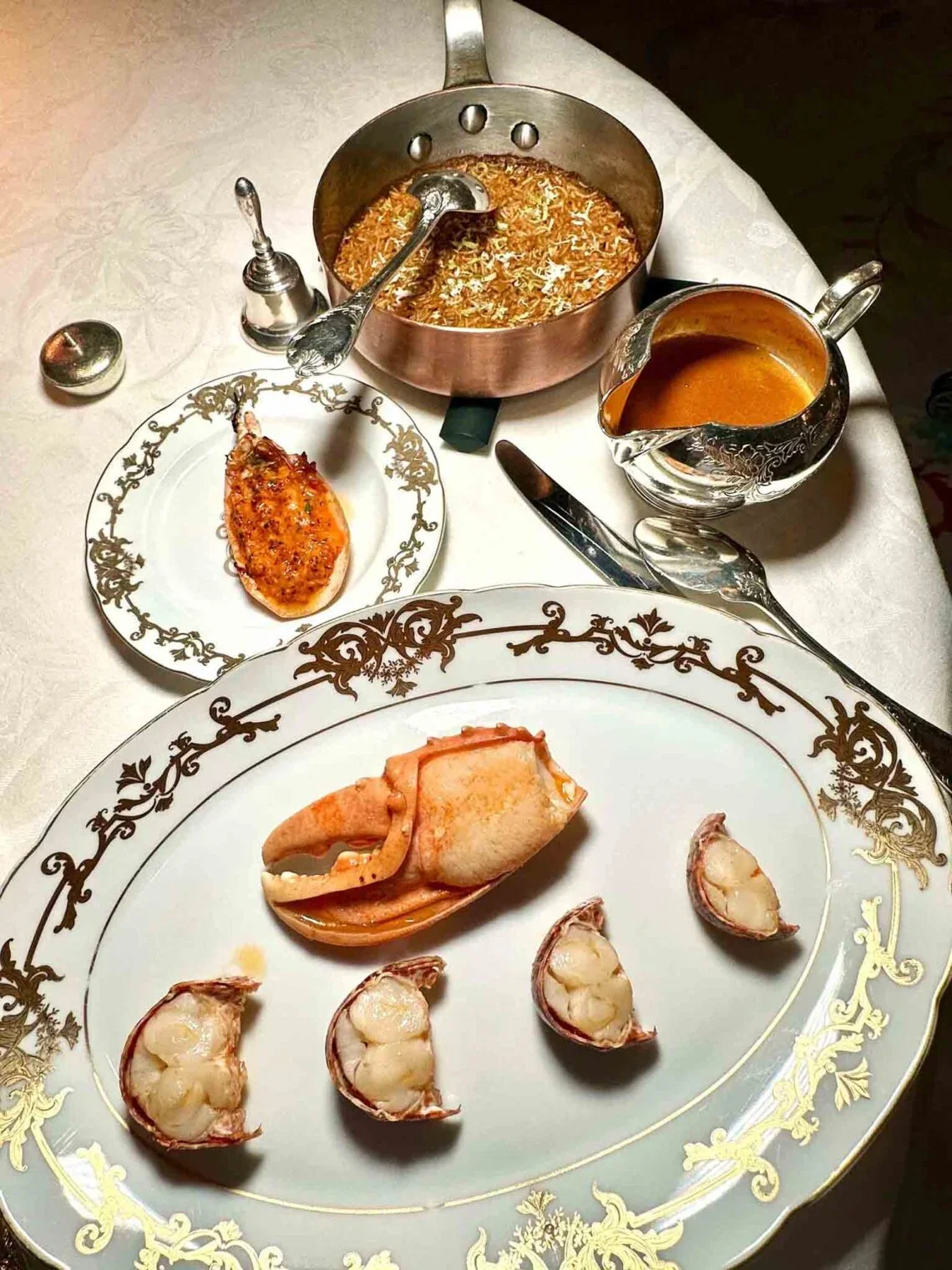 Assiette de homard coupé en morceaux, expresso au fond, une petite assiette avec une portion de viande en sauce, une sauce brune dans une cruche, une casserole avec du riz au safran, une petite assiette avec une portion de viande.