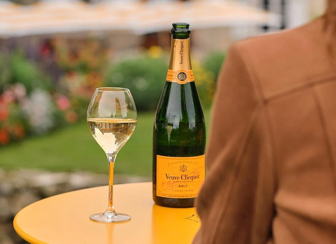 Une bouteille de champagne Veuve Clicquot et un verre de champagne sur une table jaune en extérieur, avec un fond flou de végétation.