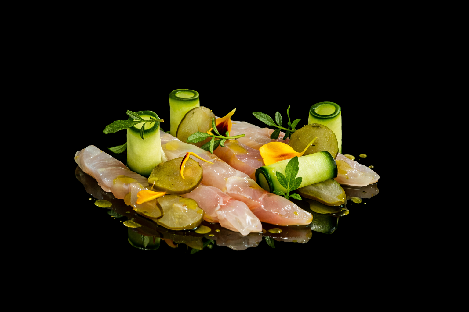 Assiette de tartare de poisson avec des rondelles de concombre, des cornichons, des feuilles de menthe, des fleurs comestibles, et un filet d'huile, sur un fond noir.