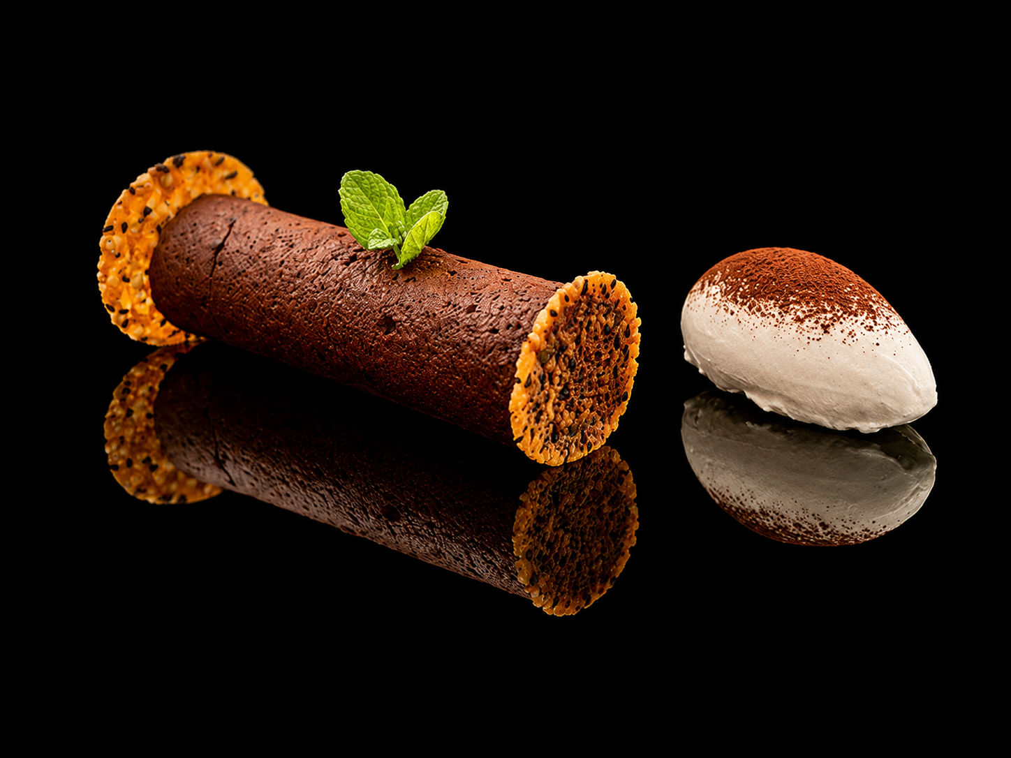 Une roulade de dessert enroulée avec une garniture croquante, décorée d'une feuille de menthe, accompagnée d'une quenelle de crème ou de mousse à la cuillère, le tout sur un fond noir reflétant la surface.