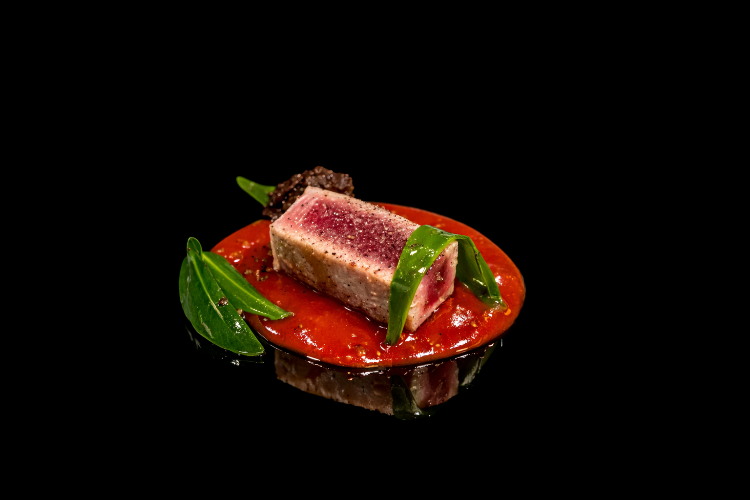 Une présentation gastronomique avec un carpaccio de thon rouge sur une assiette noire, garnie de feuilles vertes et d'une sauce rouge vif.