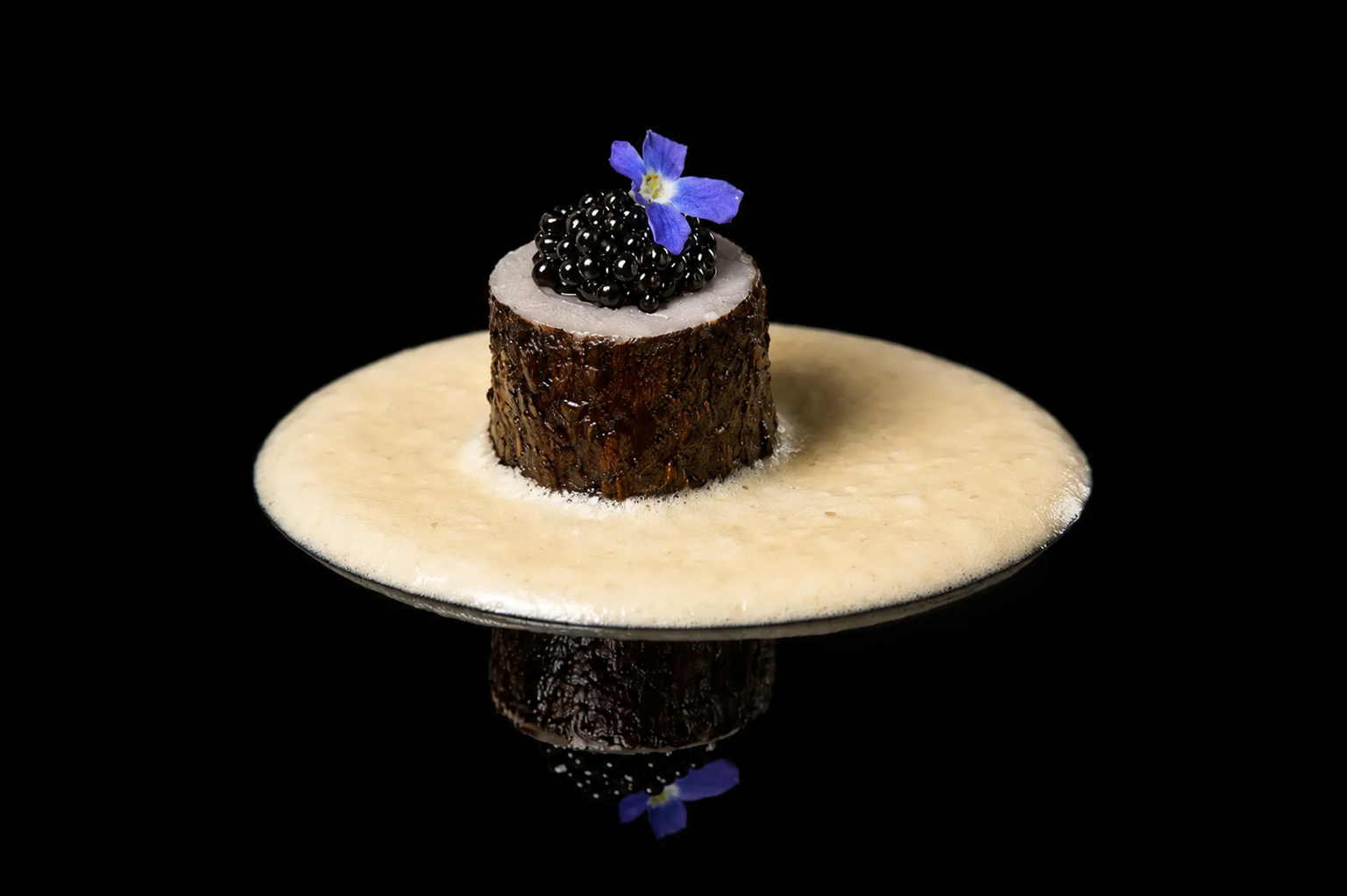 Gros plan d'une présentation culinaire avec un morceau de viande noircie, garnie de caviar noir et d'une petite fleur violette, sur une sauce beige sur fond noir.