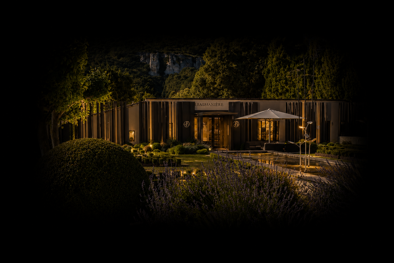 Vue nocturne d'un bâtiment moderne avec jardin et lampes d'extérieur, entouré de verdure et de montagnes en arrière-plan.