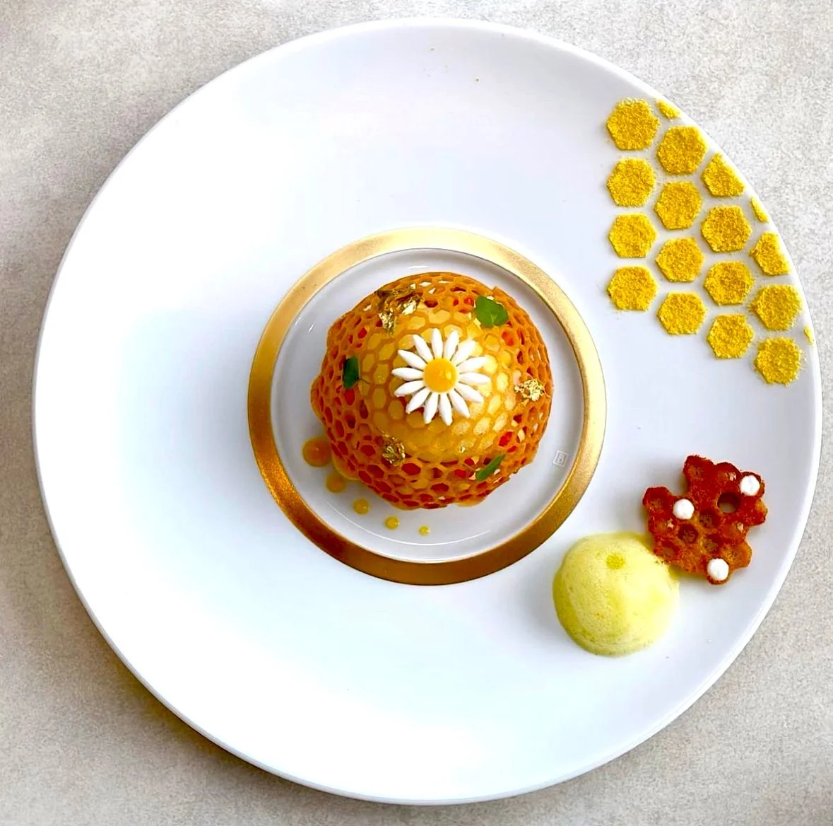 Assiette avec un dessert en forme de sphère, décorée d'une marguerite en pâte. Sur la droite, petits motifs hexagonaux jaunes, un motif rouge et un melon vert et blanc.